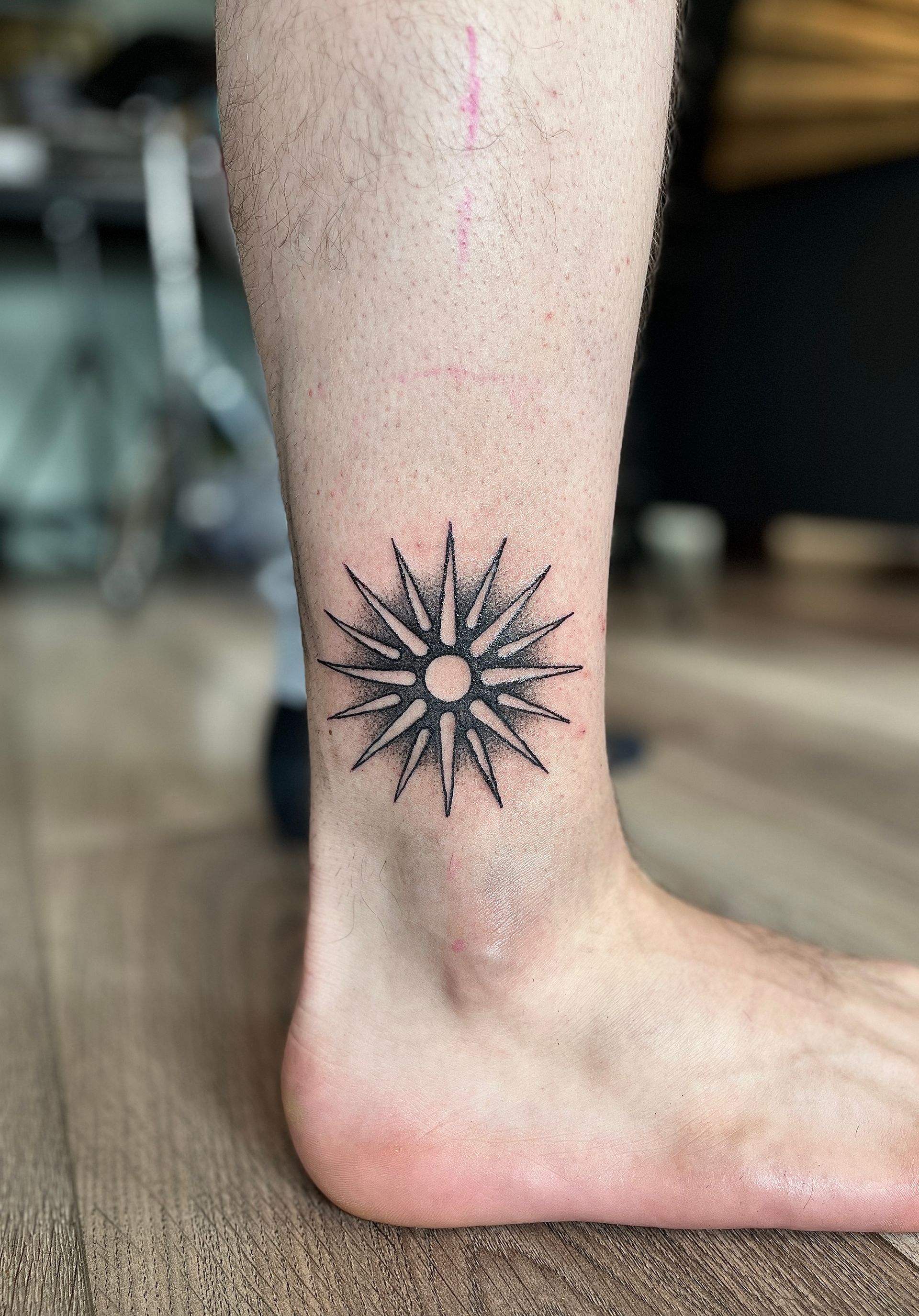 Enkeltattoo van een zwarte zonnestralen op een bloot been en een voet die op een houten vloer staat.