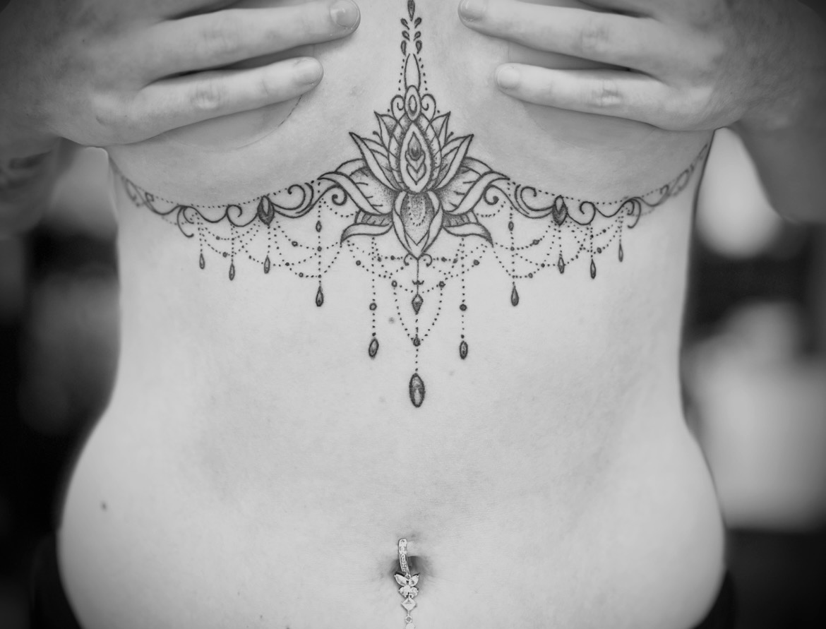 Zwart-wit close-up van een torso met een sierlijke tatoeage op de onderrug en een navelpiercing.