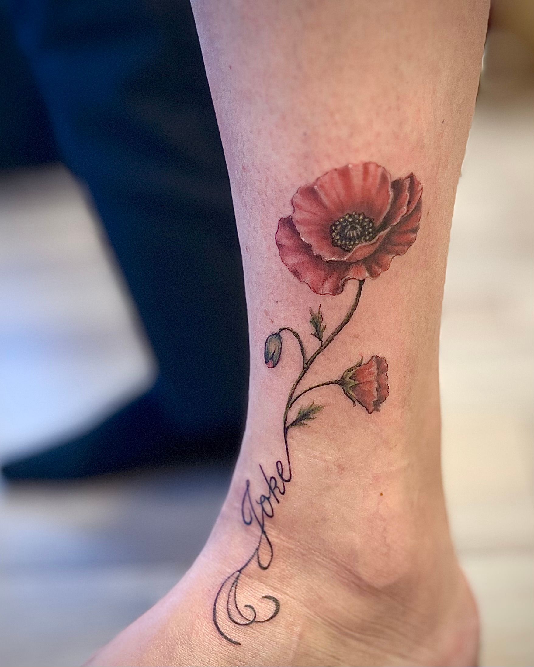 Tattoo van een rode klaproos met bladeren en sierlijke letters op het onderbeen, vlakbij de enkel.