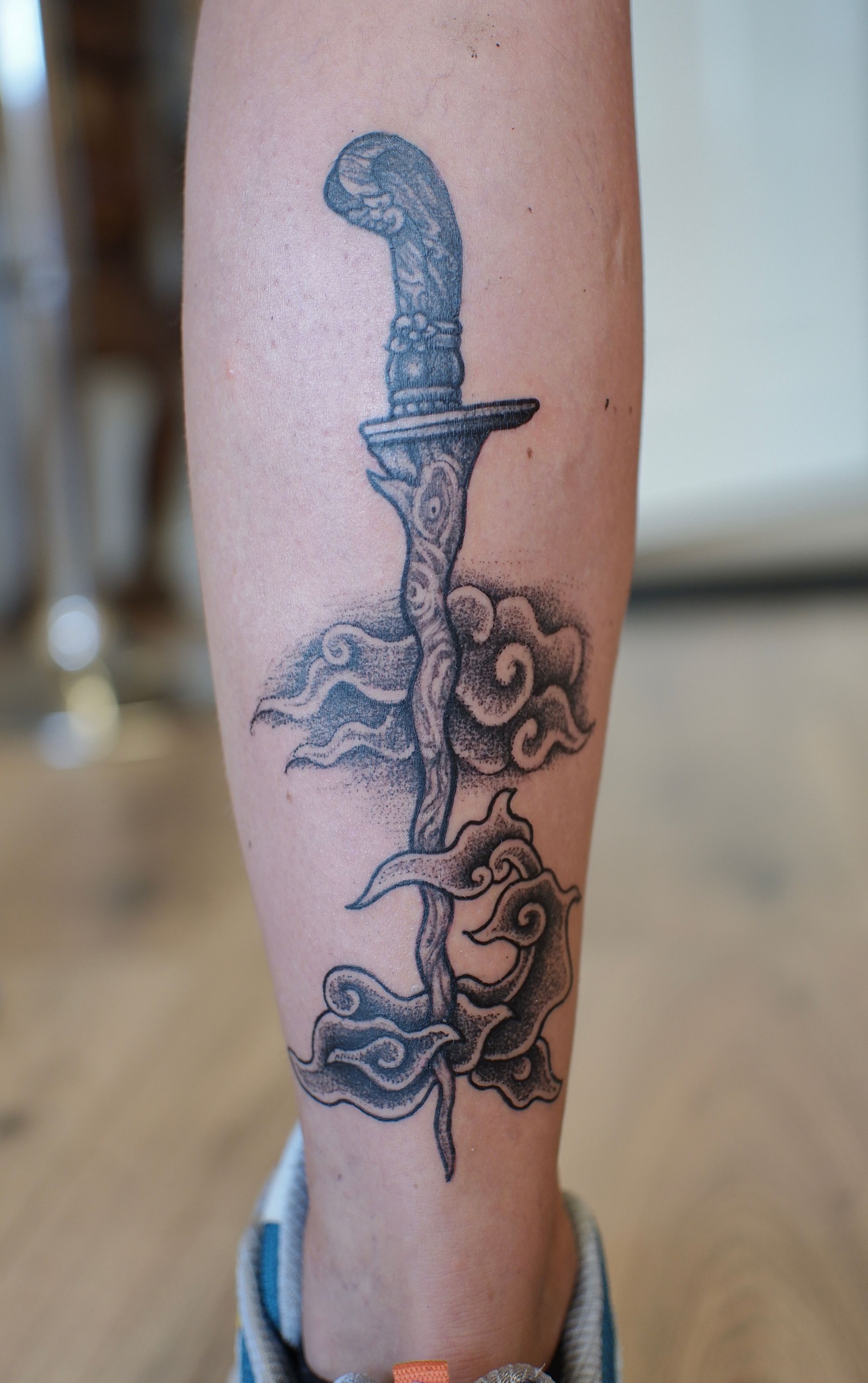 Zwart-grijze tatoeage van een dolk op een onderbeen, met bloemen en een slang eromheen gewikkeld.