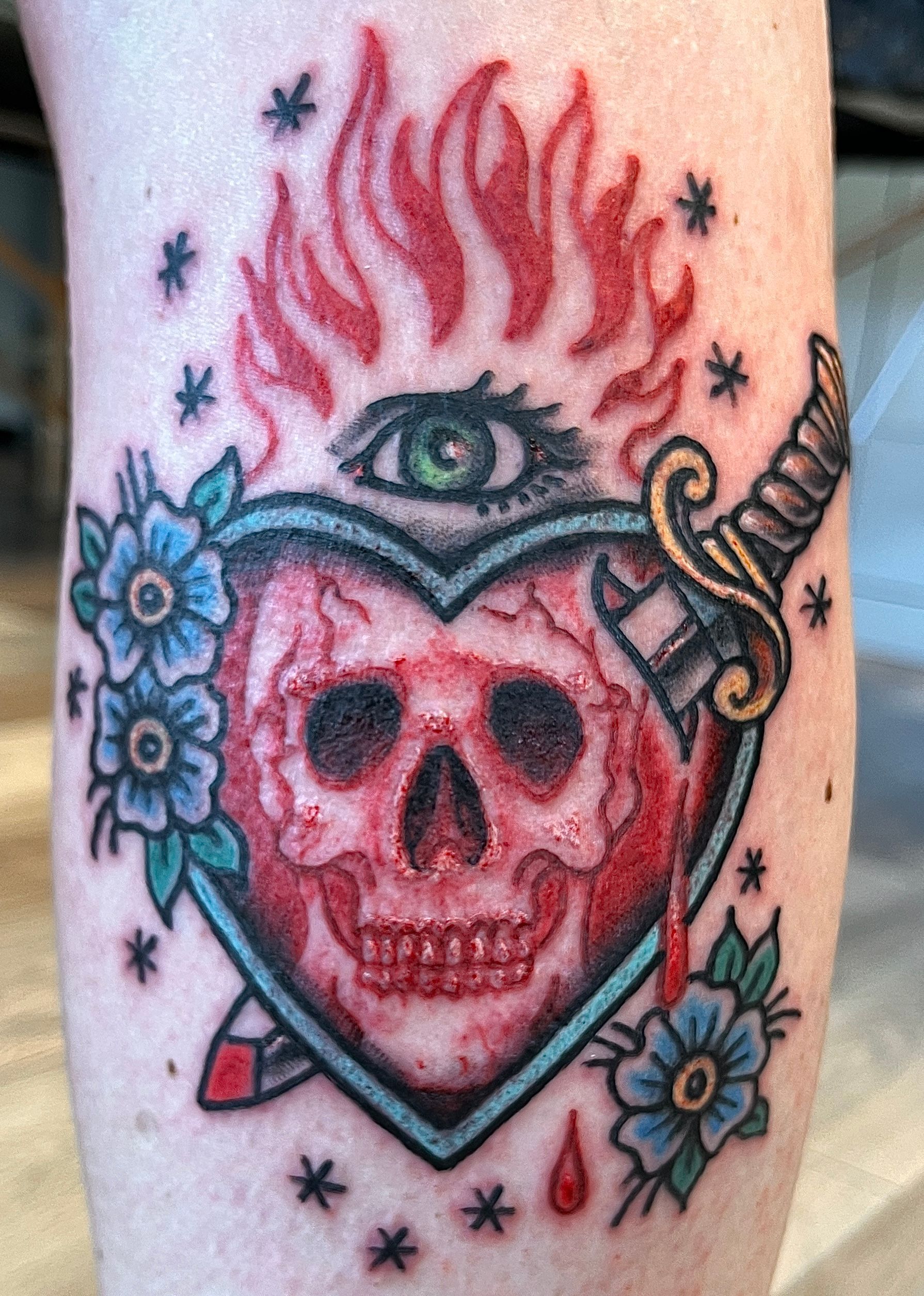 Tattoo van een vlammende schedel in een hart, met blauwe bloemen en sterren op de huid.