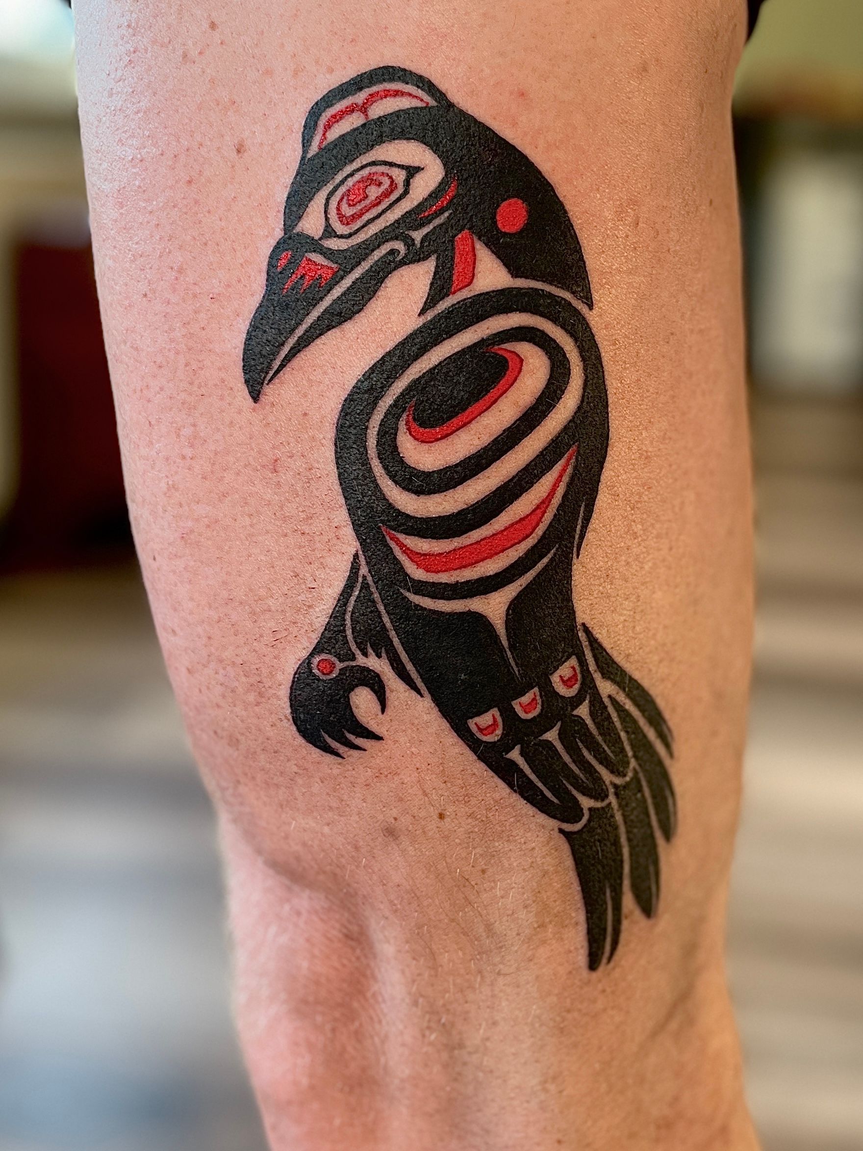 Een gestileerde vogeltattoo in zwart en rood op het been, met cirkelvormige patronen en een scherpe snavel.