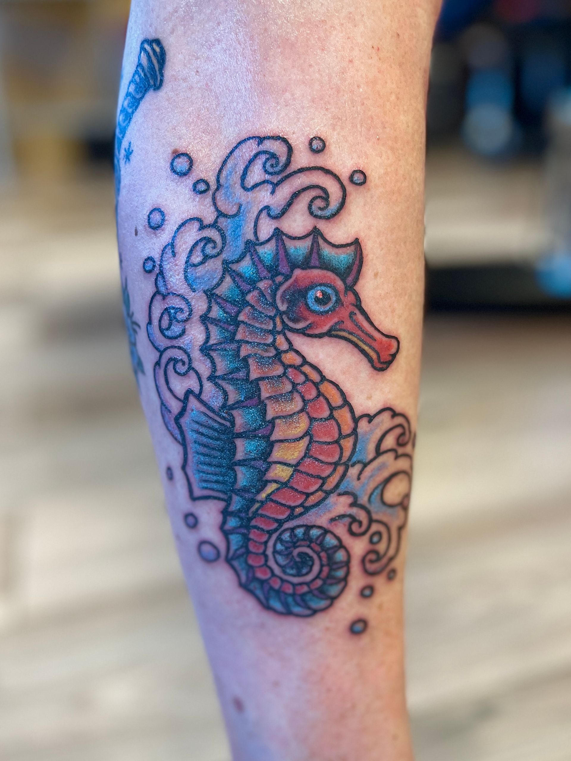 Tattoo van een zeepaardje met blauwe en rode lijnen en wervelende decoratieve accenten op de kuit.