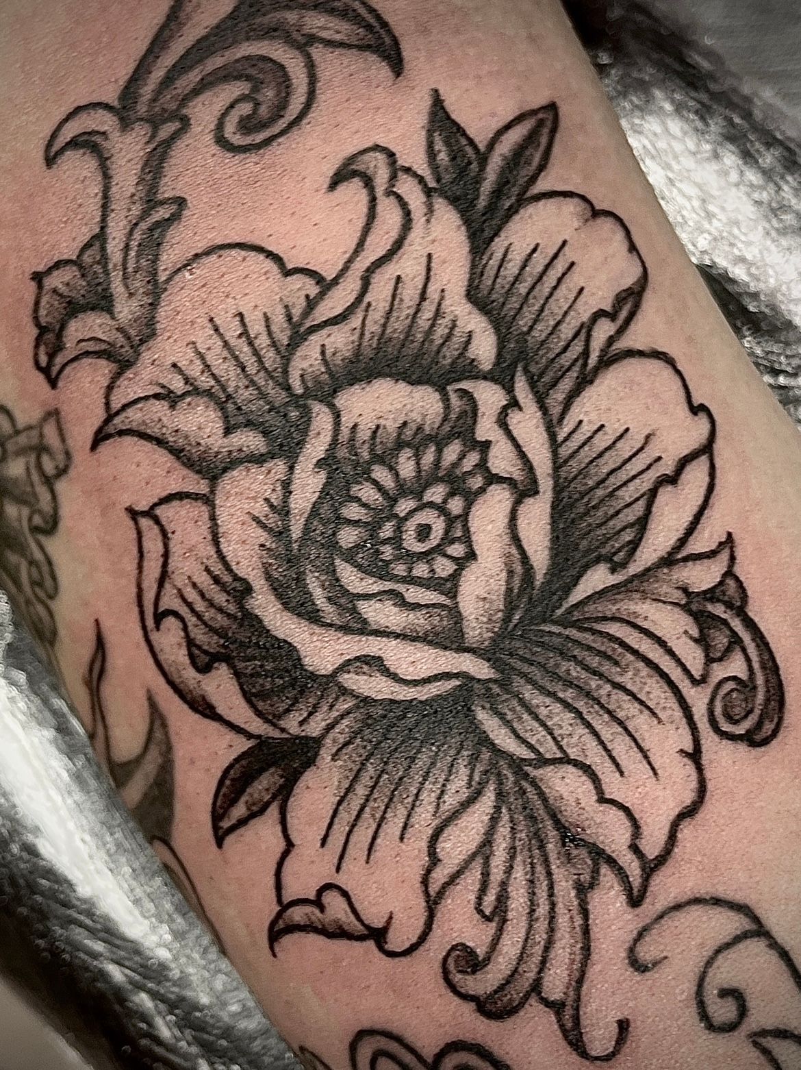 Zwart-grijze bloementattoo van een grote roos met decoratieve bladeren en krullen op de huid.