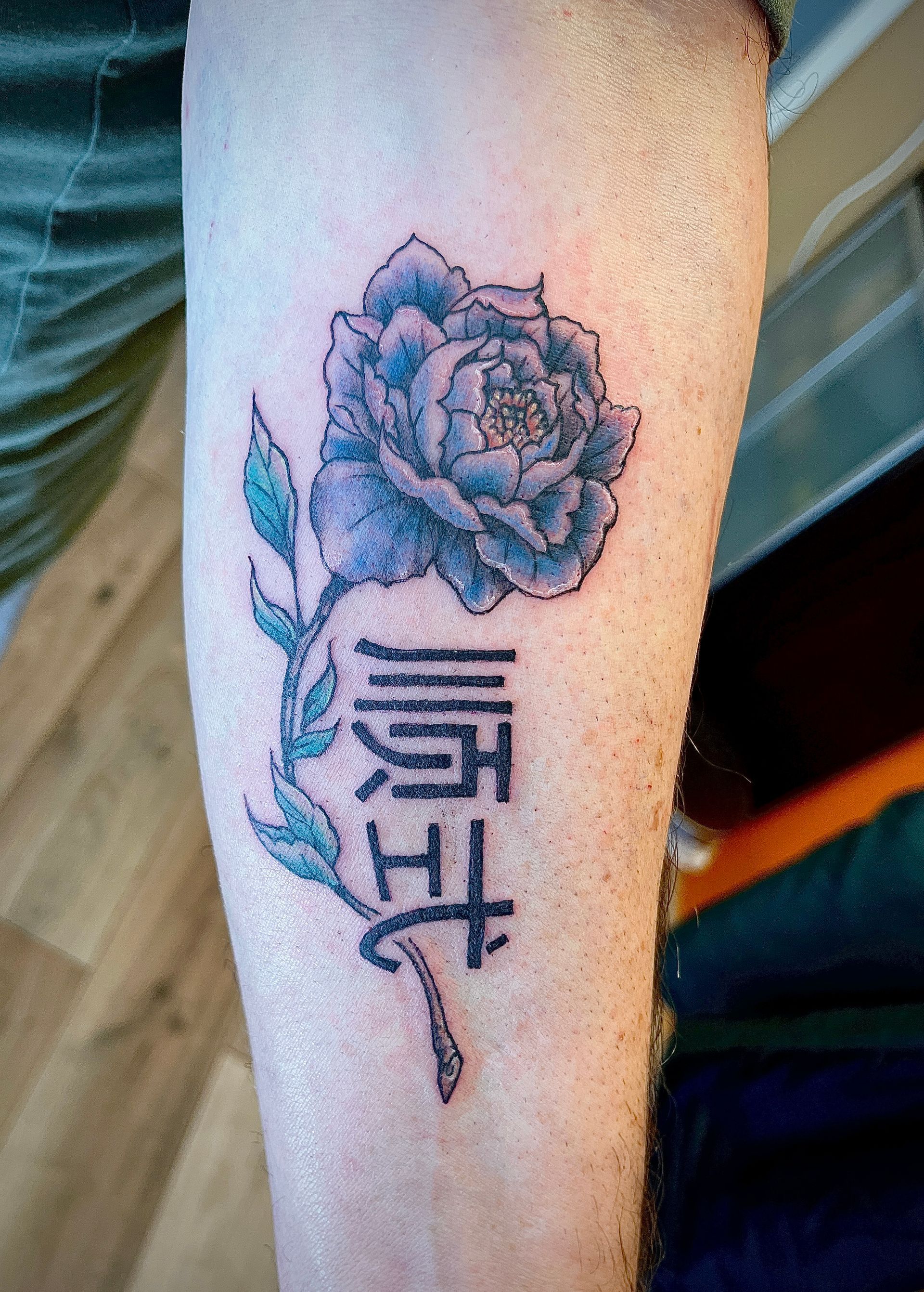 Tattoo op de onderarm van een roos boven verticale, gestileerde letters in zwarte inkt op een lichte huid.