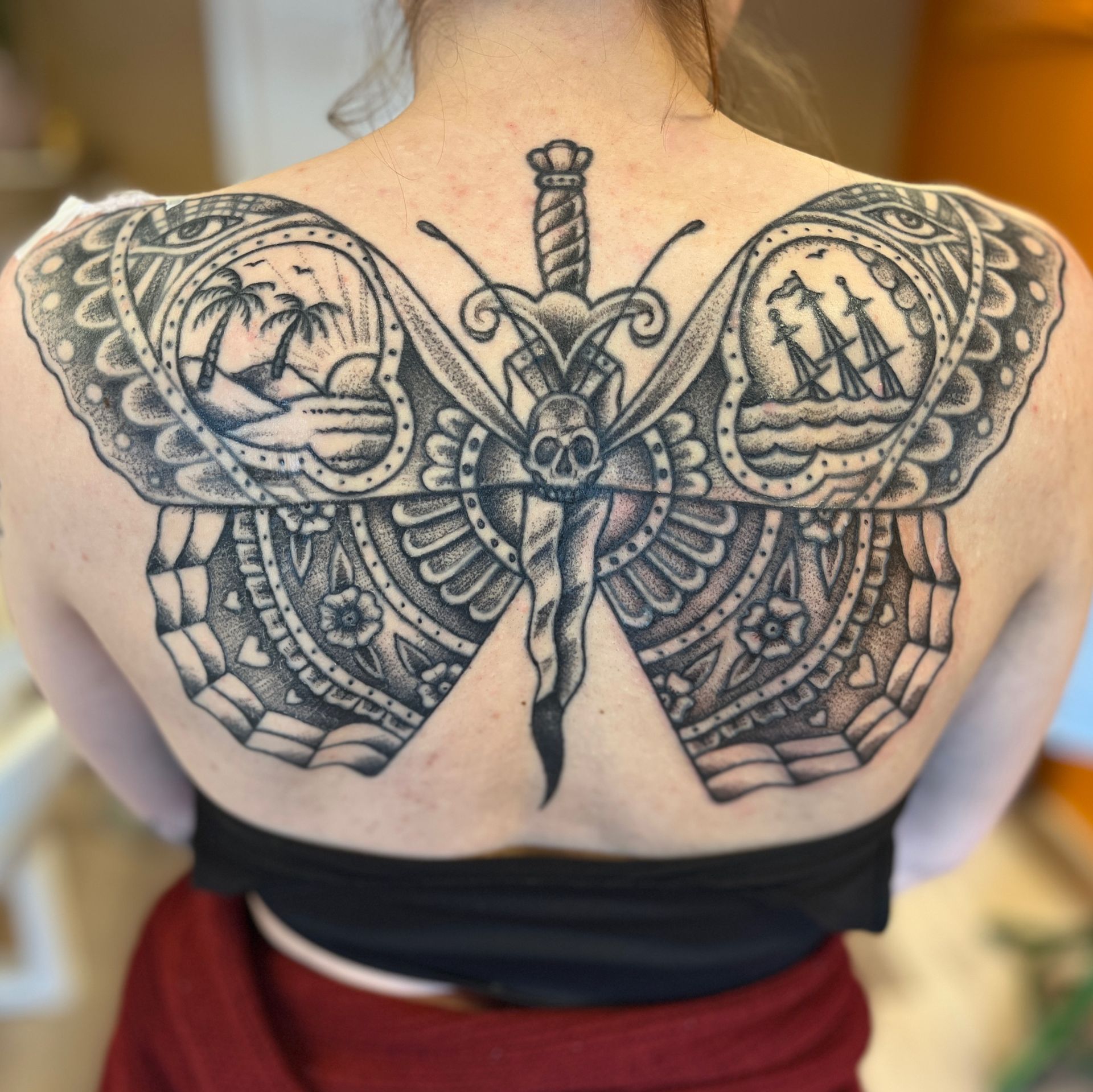 Tattoo op de rug van een gedetailleerde zwarte vlinder met mandala-vleugels en cirkelvormige natuurtaferelen.