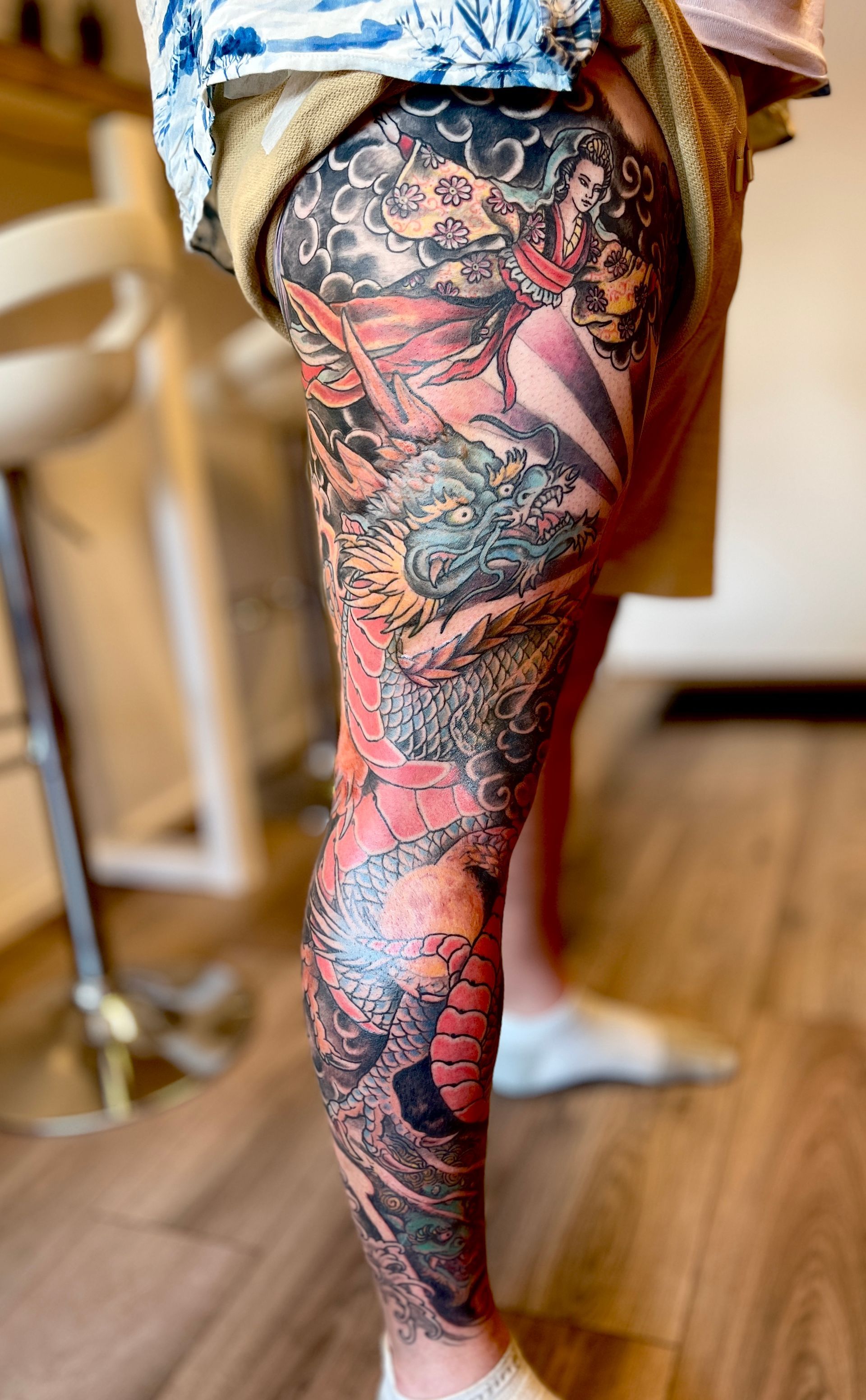 Het been van een persoon met een kleurrijke tatoeage op de arm, staand binnenshuis op een houten vloer met een kruk in de buurt.