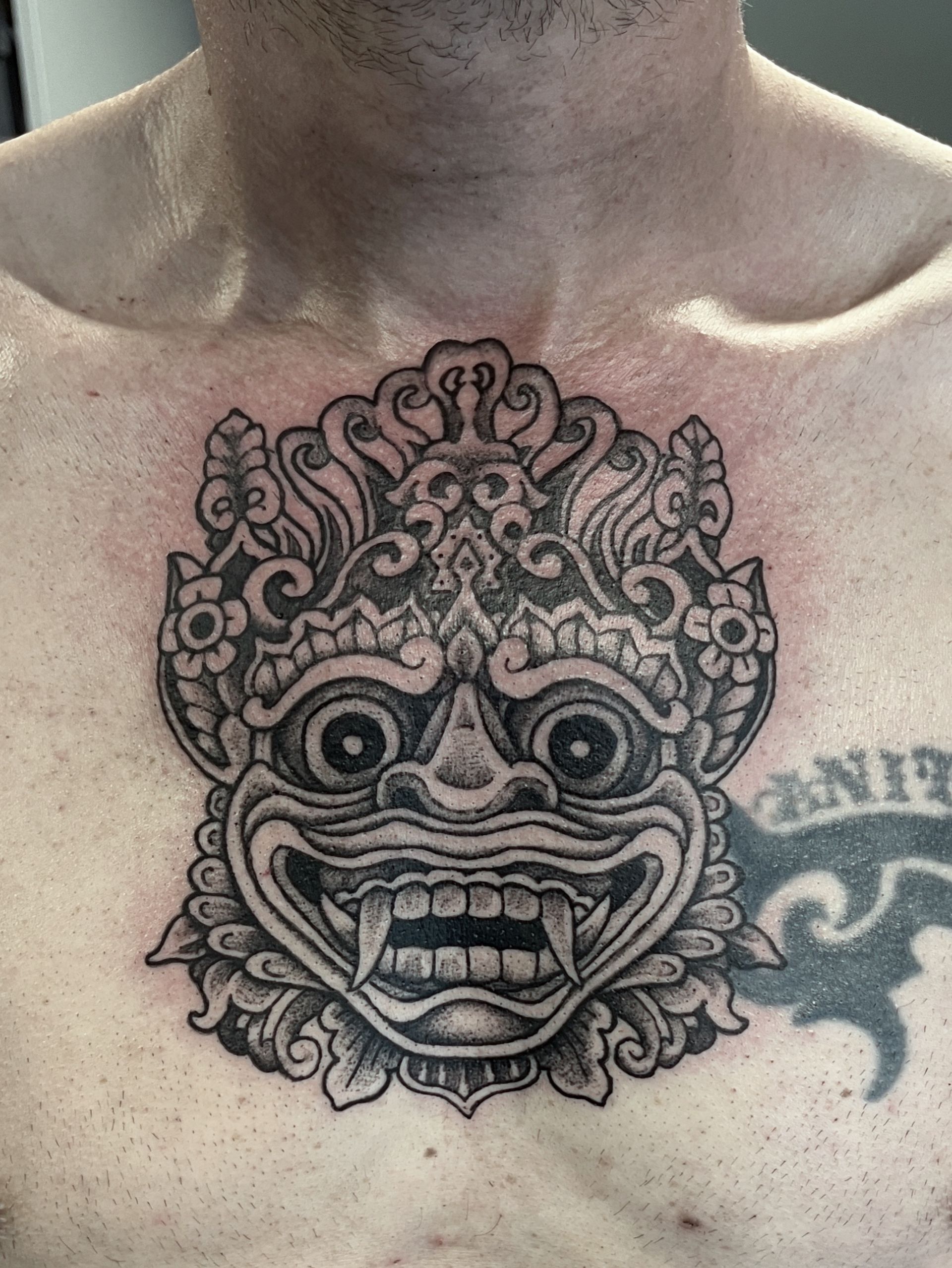 Borst met een zwart-grijze, sierlijke maskertattoo met een gezicht met grote ogen en ingewikkelde, wervelende details.