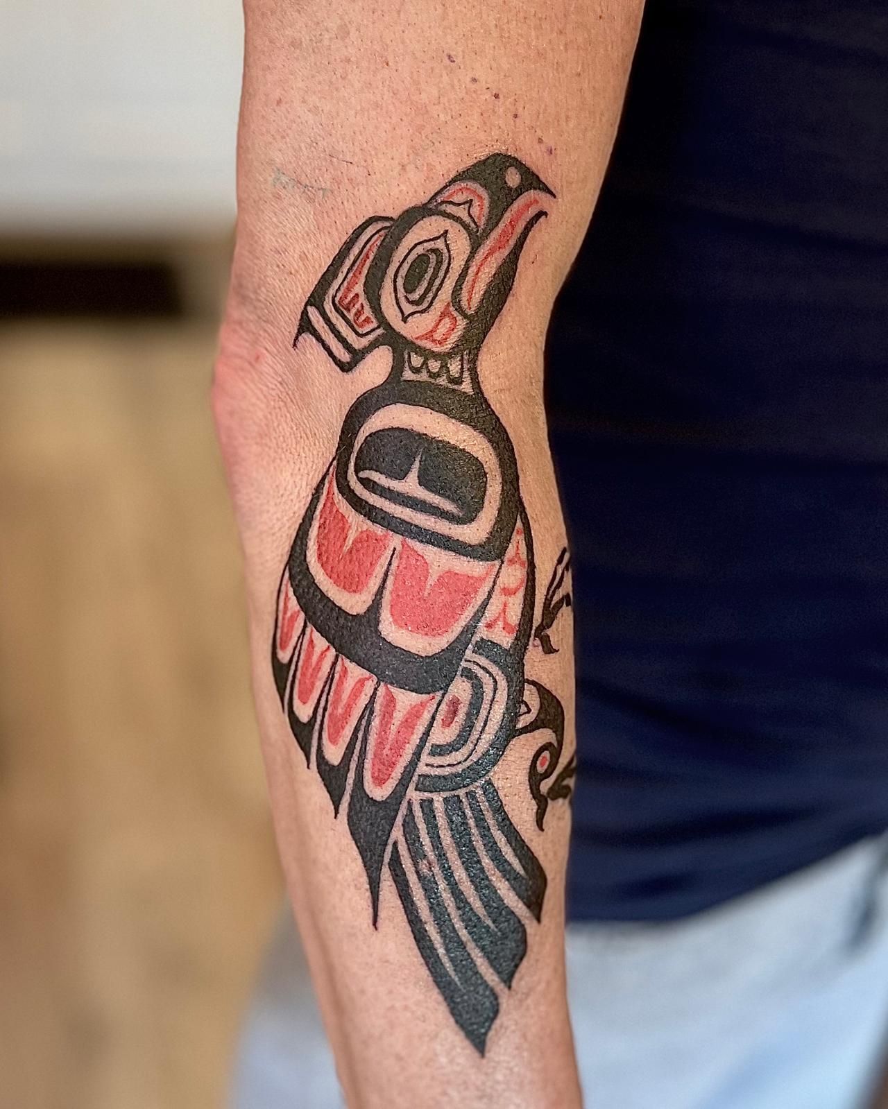 Close-up van een kleurrijke vogeltattoo op de arm van een persoon, afgebeeld tegen een donker shirt en een lichte korte broek.