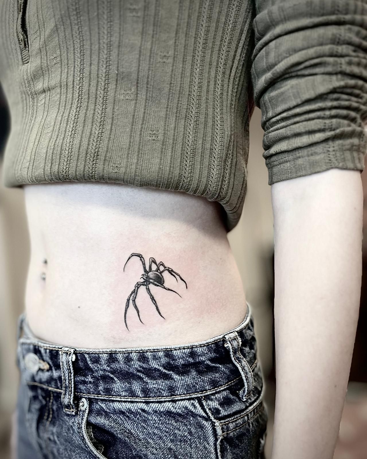 Close-up van een torso met een zwarte spinnentattoo aan de zijkant van de taille, boven de spijkerbroek.