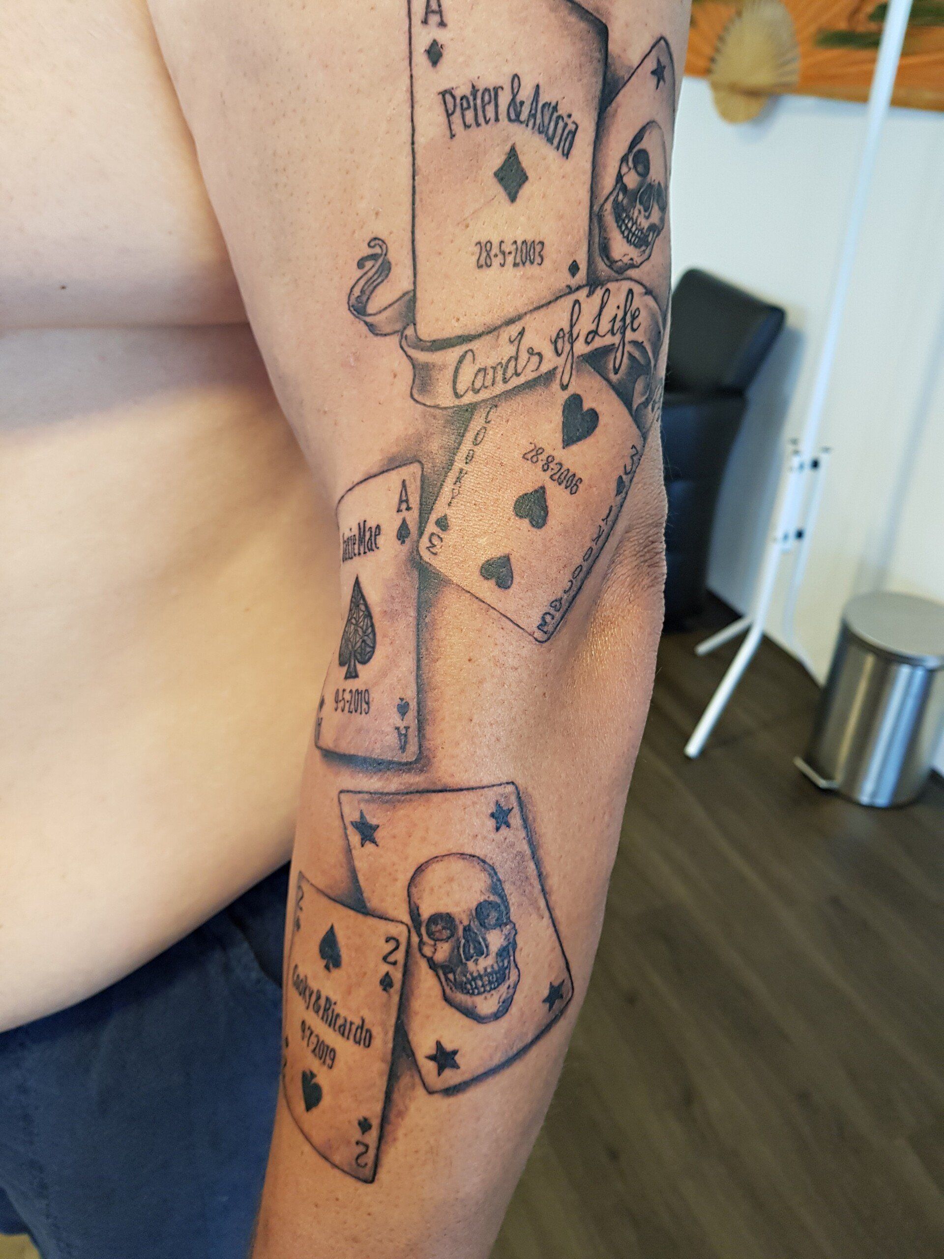 Arm met tatoeages van speelkaarten, waaronder aas en vrouw, in een informele binnenomgeving.