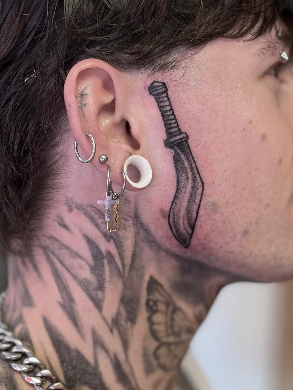 Close-up van een persoon in zijprofiel met meerdere oorpiercings en een handtatoeage achter het oor.