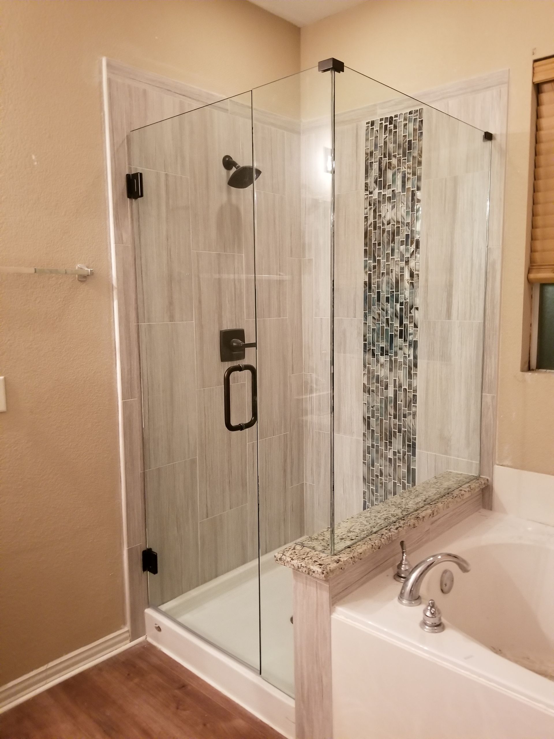 Glass corner shower enclosure beside a white bathtub in a beige bathroom with granite trim