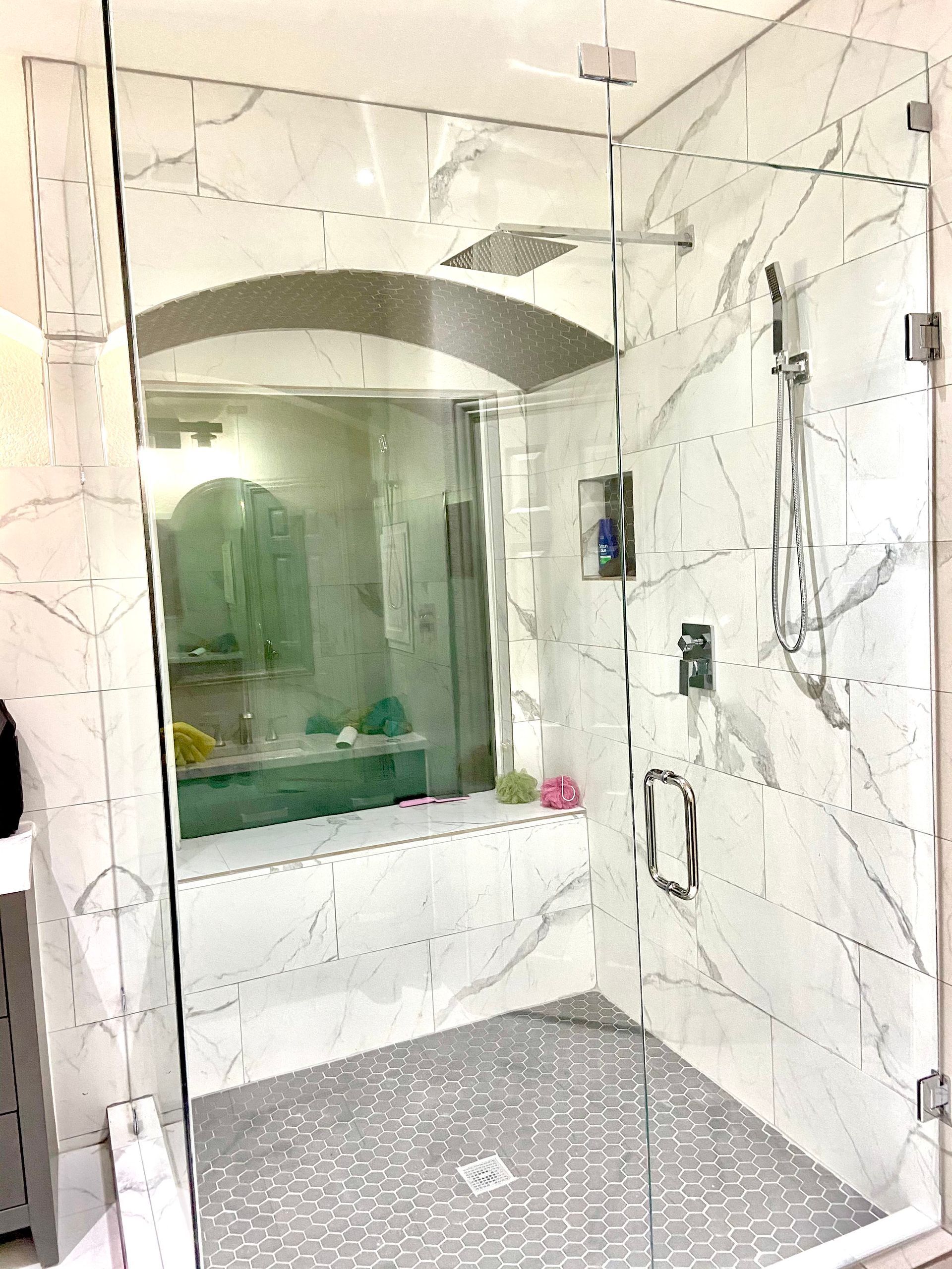 Glass-enclosed marble shower with bench, arched mirror, and gray pebble floor in a bathroom