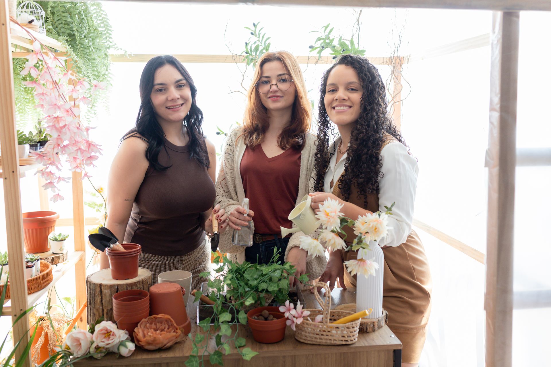 Três mulheres estão de pé, uma ao lado da outra, em frente a uma mesa cheia de plantas em vasos.
