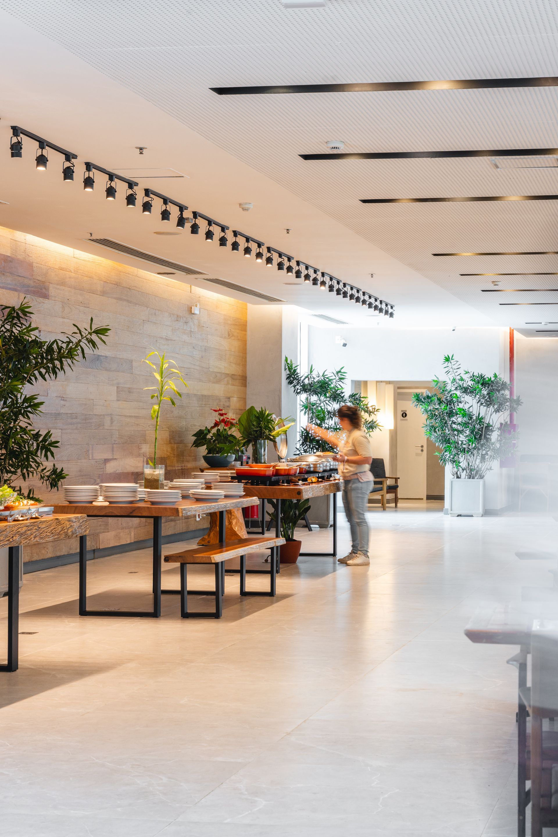 Uma pessoa está perto de uma mesa de bufê em um ambiente interno, em uma área de jantar moderna com plantas e iluminação de trilhos.