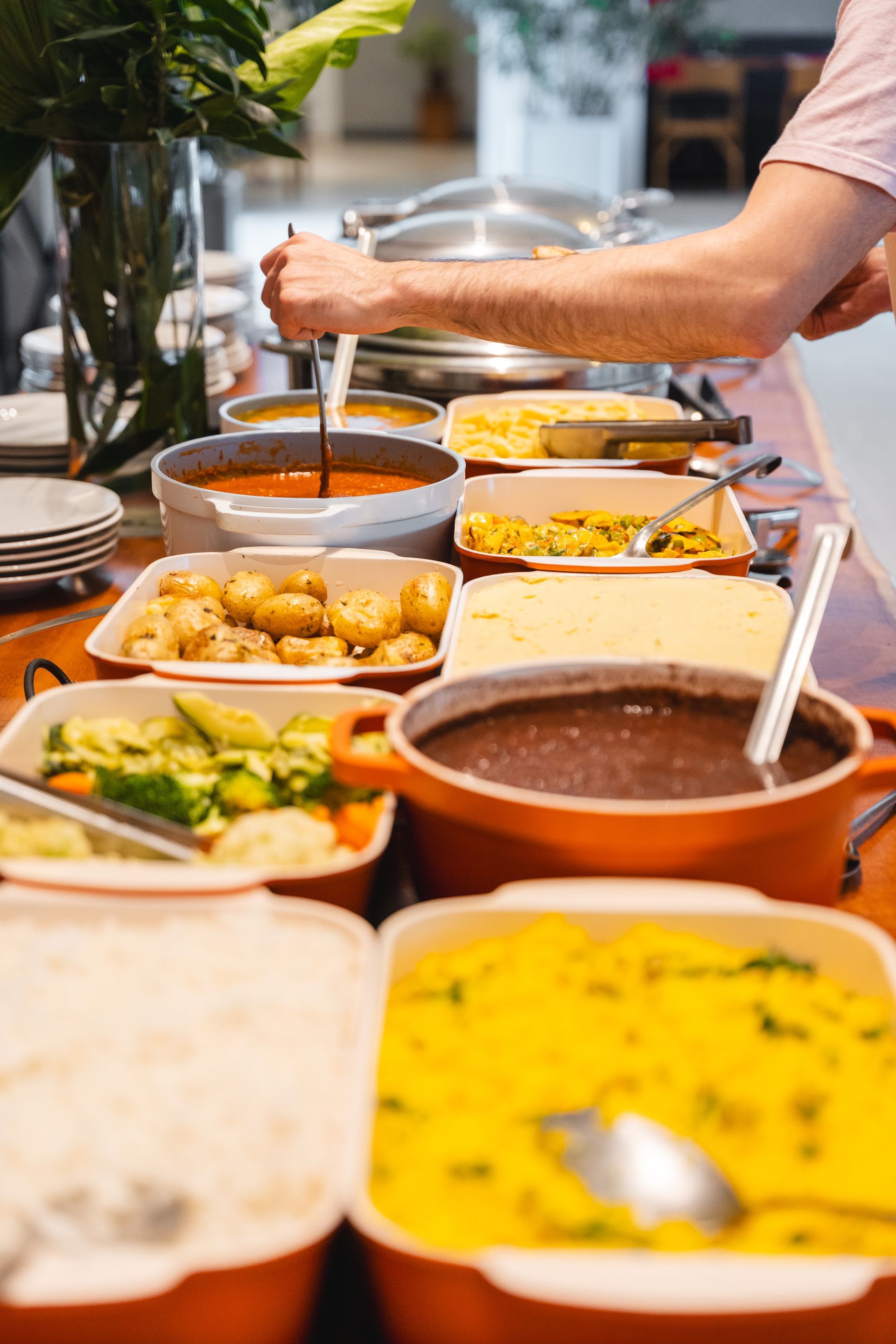 Uma mesa de bufê com vários pratos; uma pessoa serve um ensopado. Comida colorida em pratos laranja e brancos.