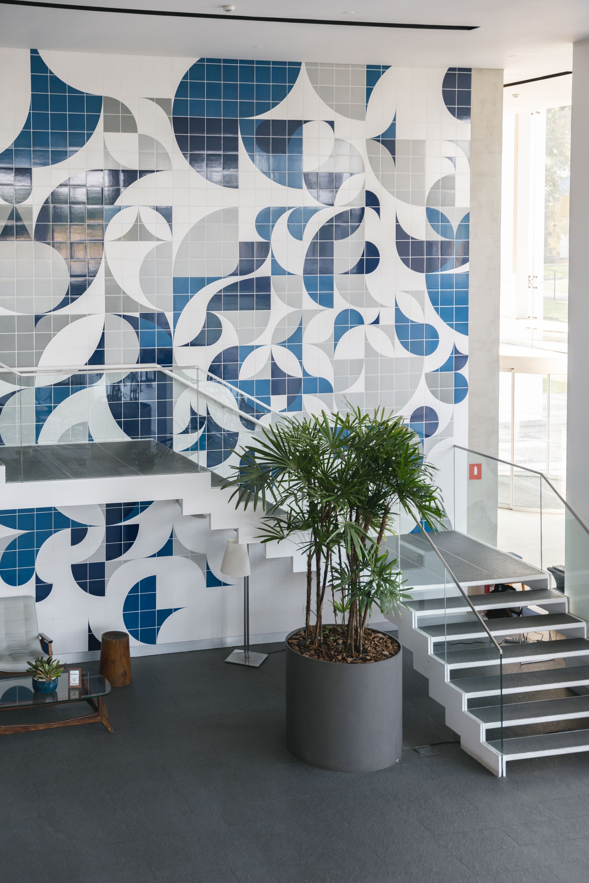 Um saguão moderno com uma parede estampada em azul e branco. Uma escada leva ao andar superior, com um vaso de plantas em primeiro plano.