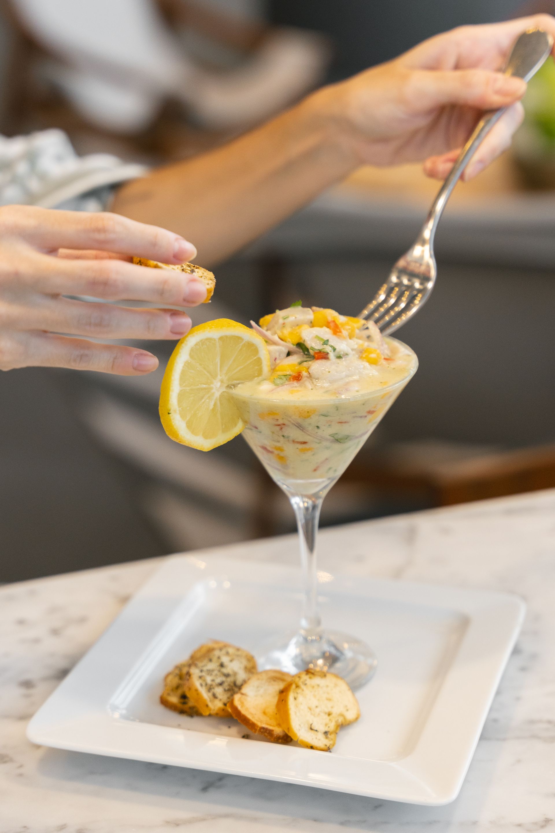 Pessoa decorando um aperitivo cremoso servido em uma taça de martini com limão e crostini em um prato branco.