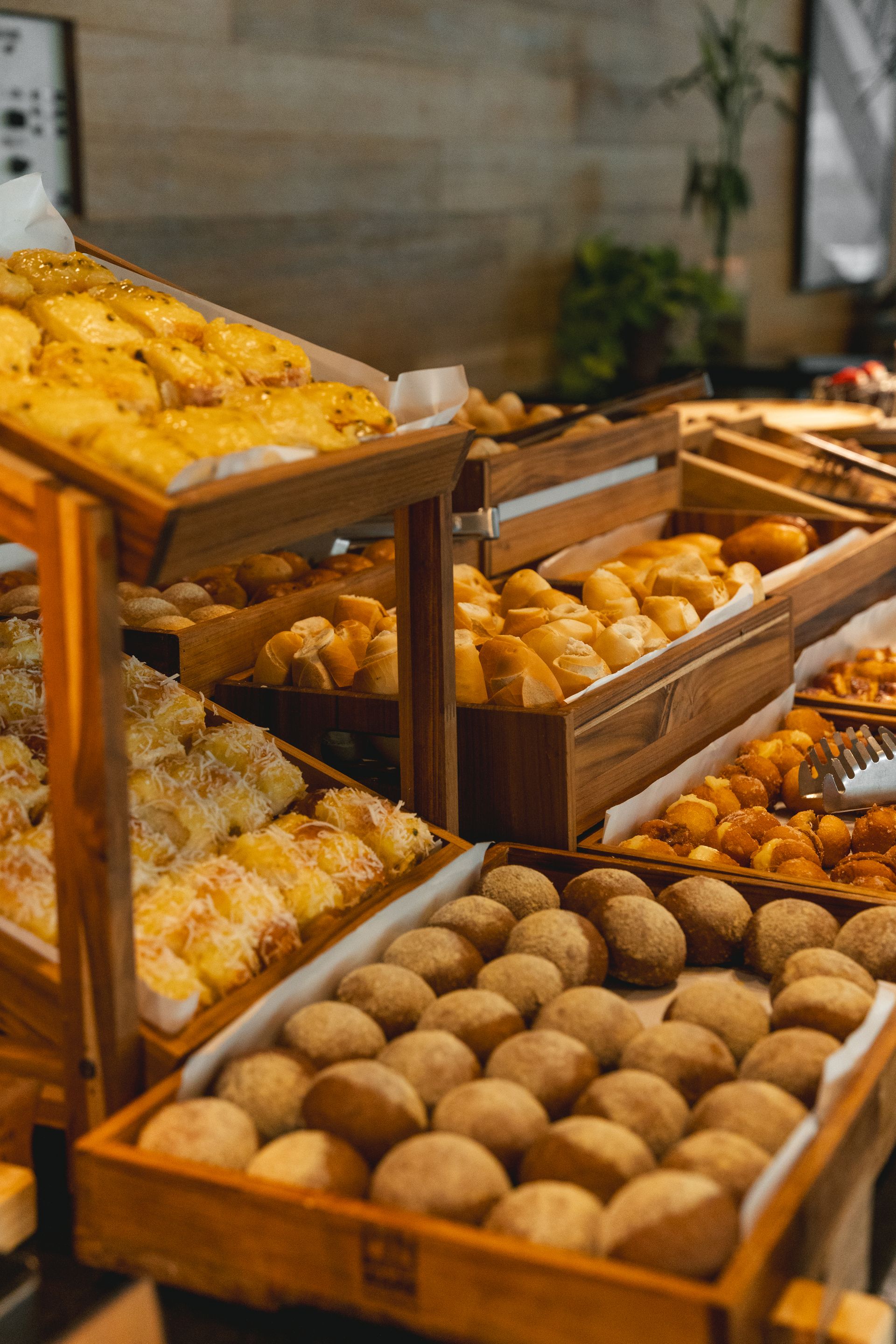 Expositor de buffet de madeira com diversos produtos de panificação, incluindo pãezinhos e doces.