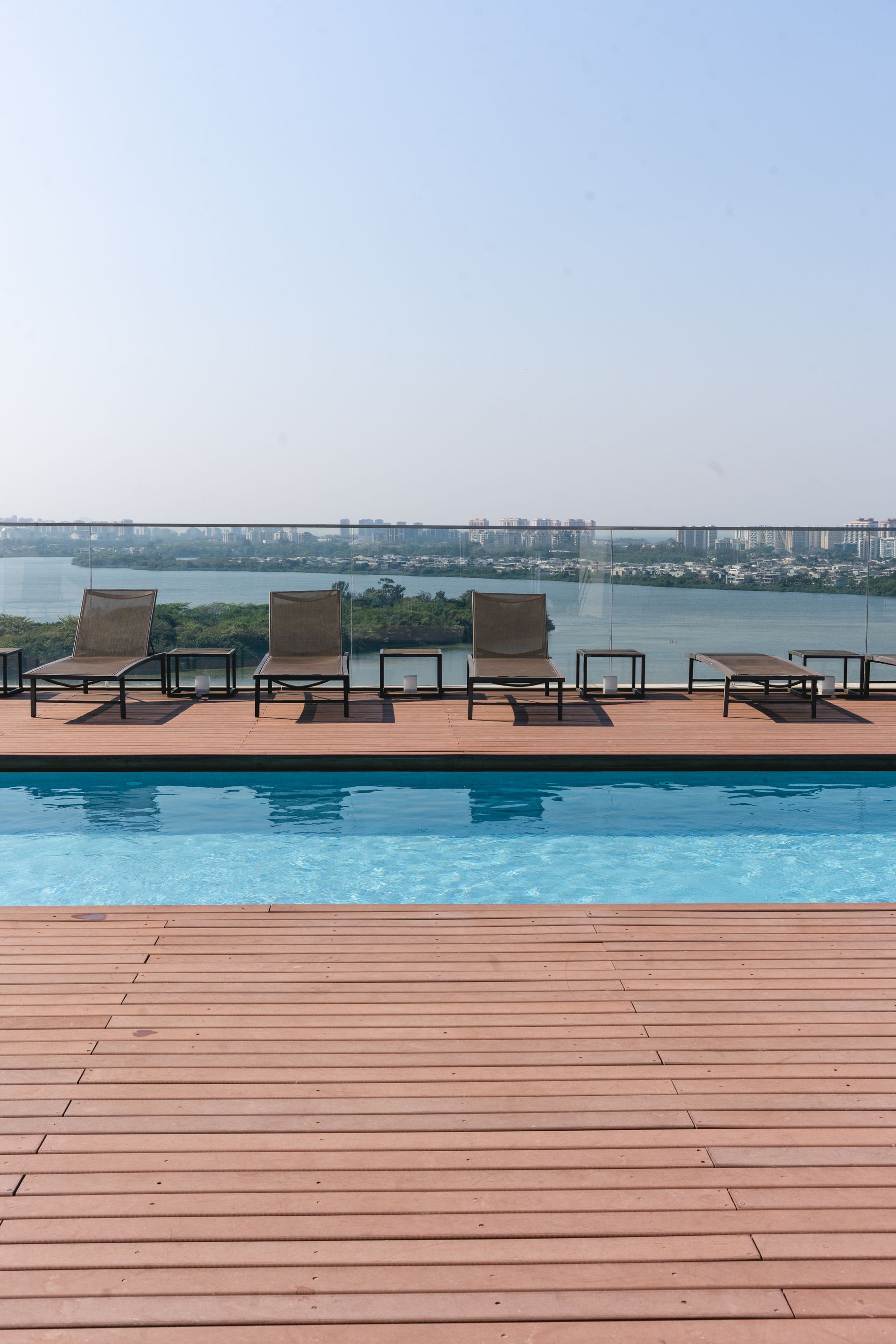 Piscina com deck de madeira e espreguiçadeiras com vista para o rio e a paisagem urbana sob um céu claro e nublado.