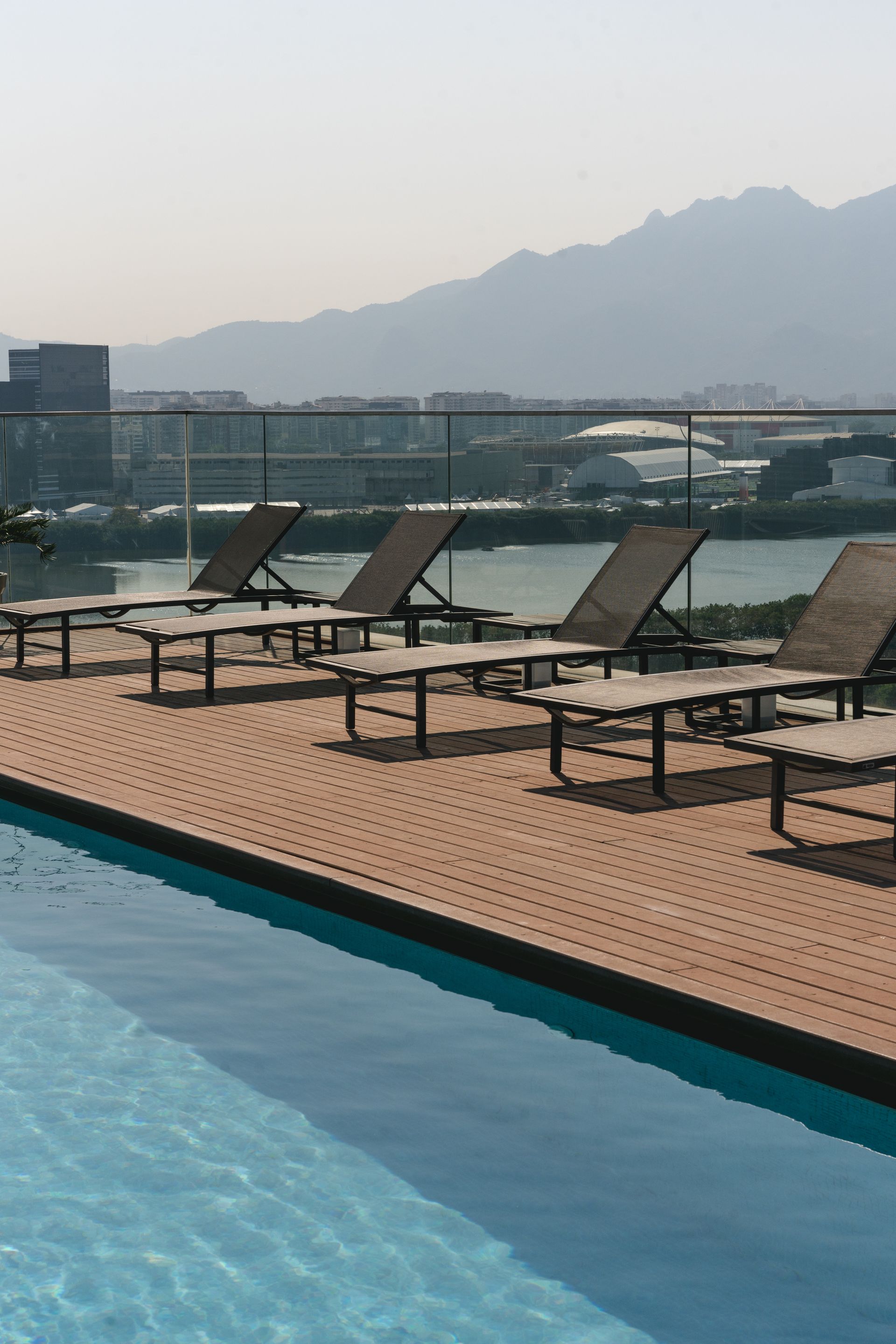 Vista da piscina: espreguiçadeiras vazias de frente para um corpo d'água e montanhas distantes.