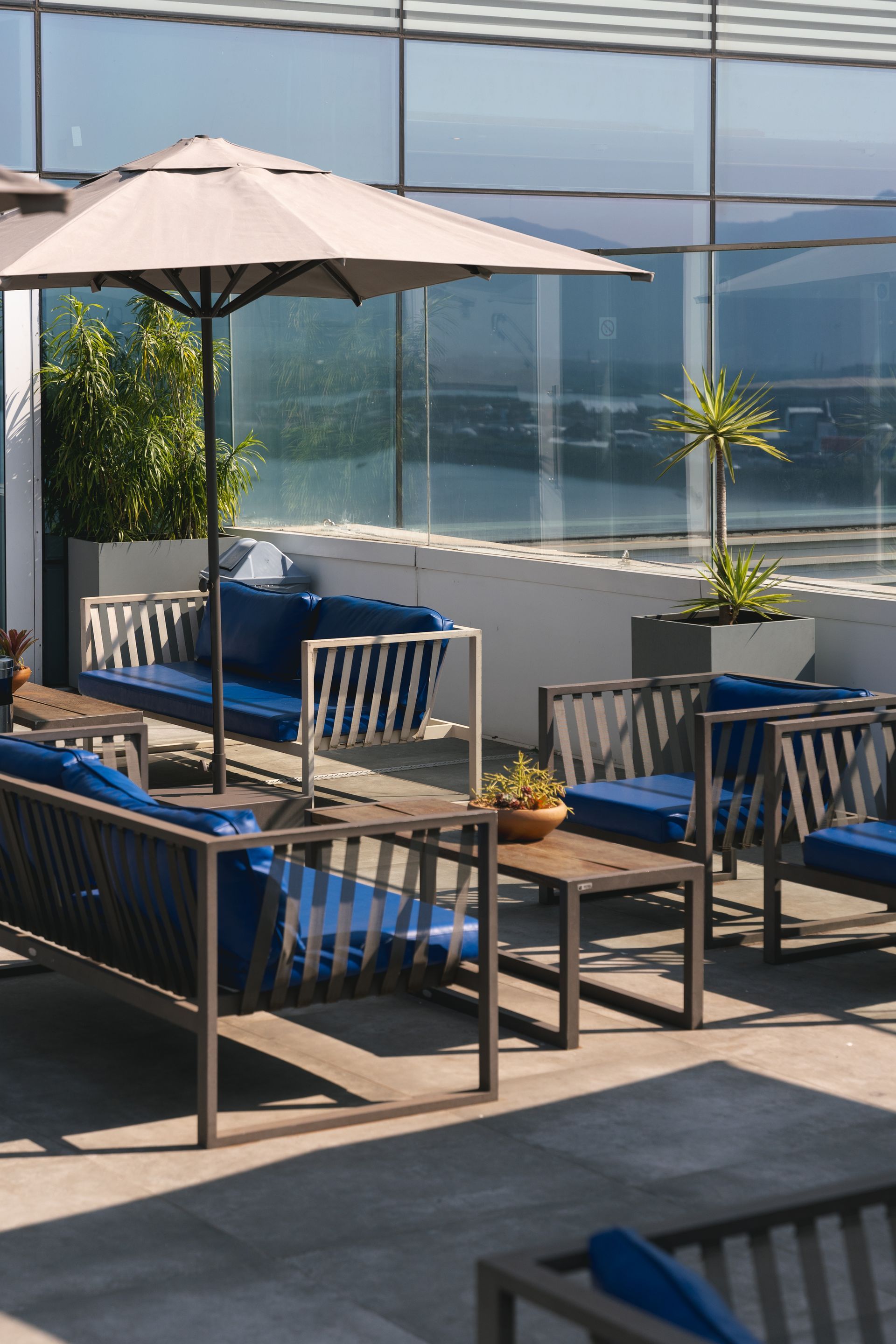 Terraço com assentos almofadados azuis, mesas pequenas e um guarda-sol bege. Plantas em vasos, com uma paisagem urbana visível ao fundo.