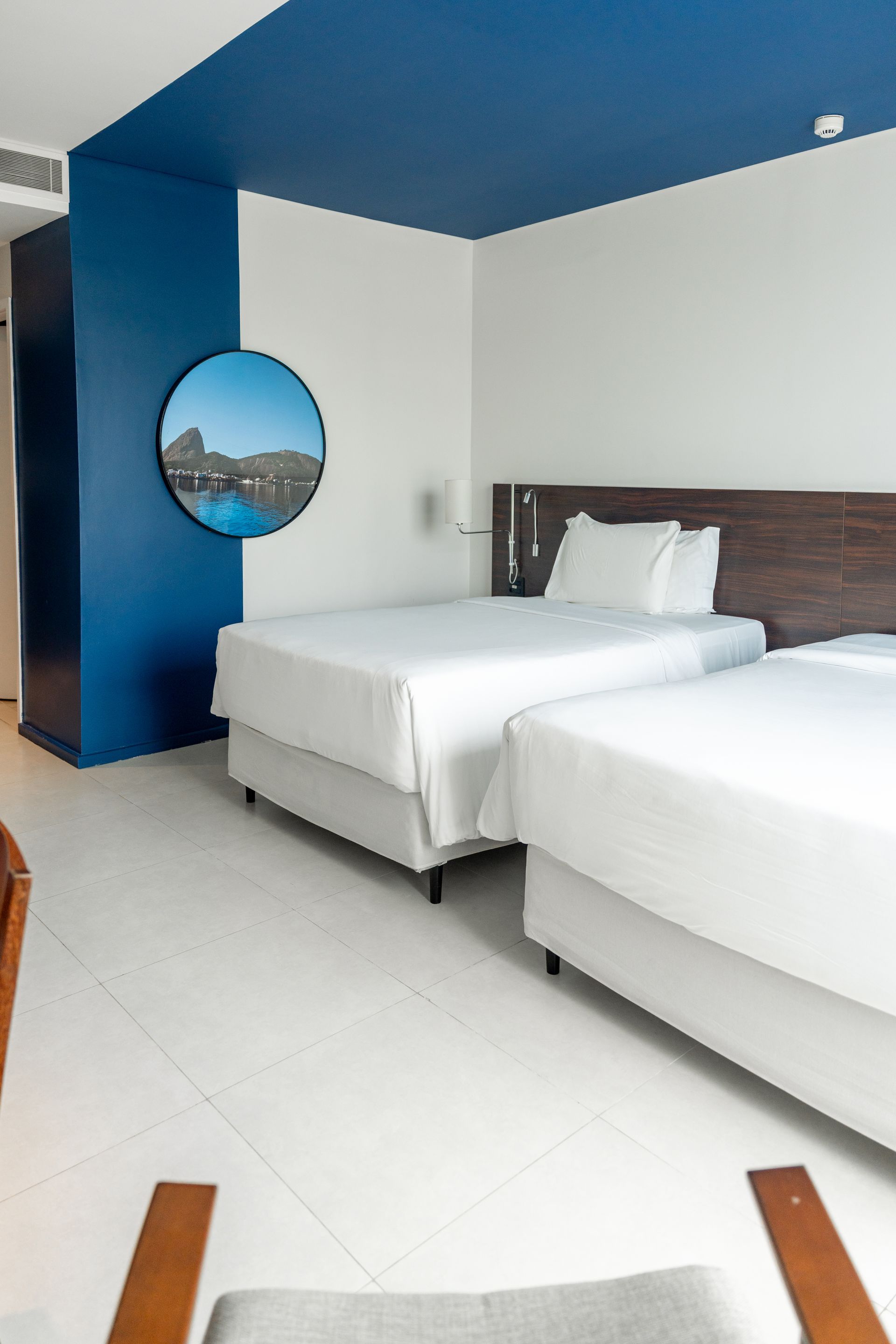 Um quarto de hotel com duas camas de solteiro cobertas com roupa de cama branca. A parede tem um esquema de cores azul e branco, com uma obra de arte redonda.