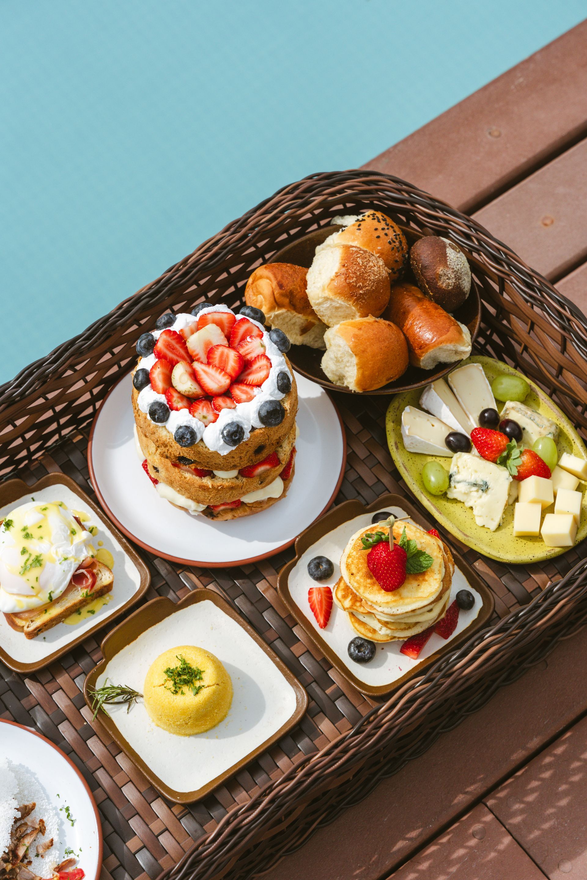 Uma bandeja de brunch com comida: bolo com frutas vermelhas, panquecas, pãezinhos, ovos Benedict, frutas, queijo, à beira de uma piscina.