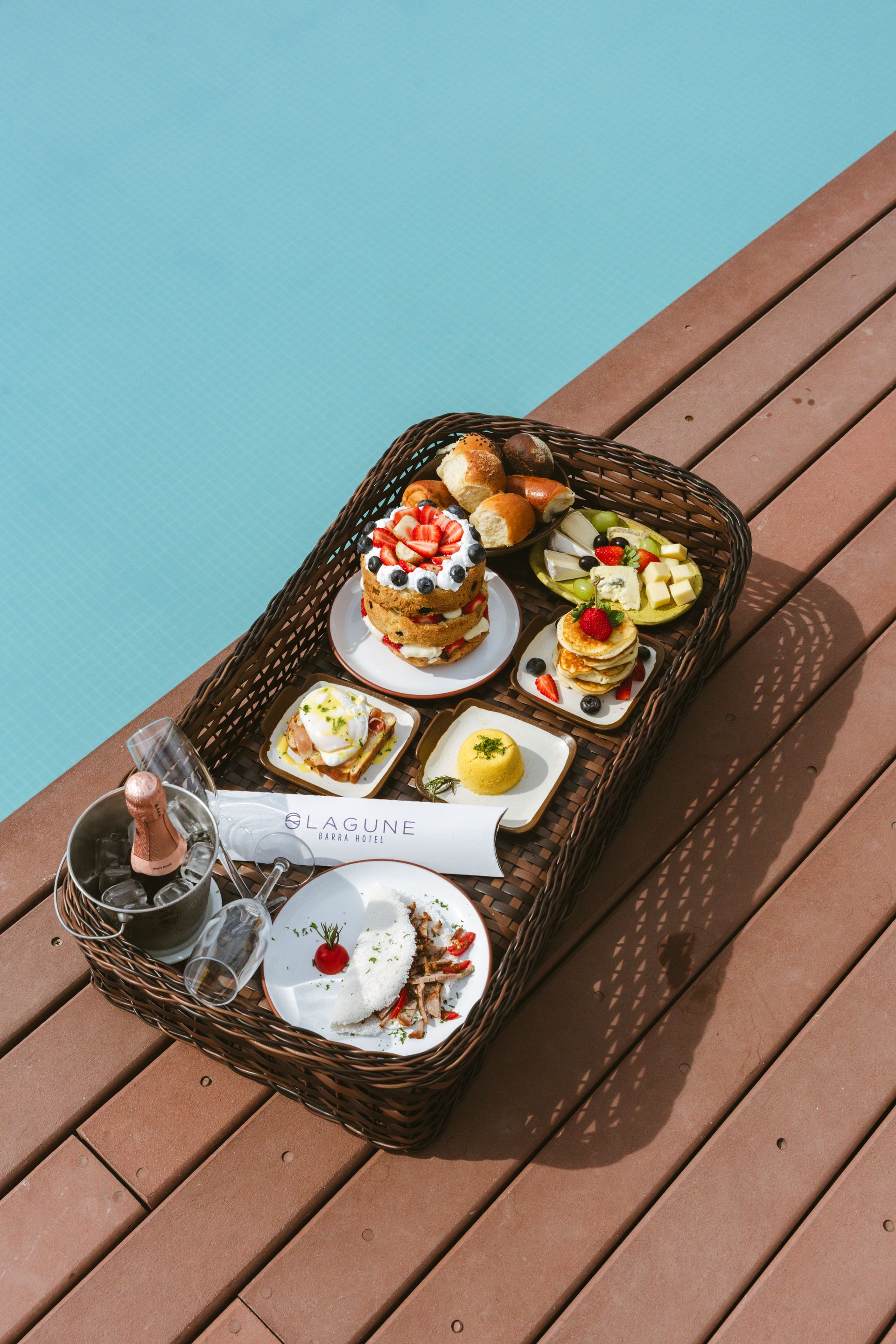 Bandeja de café da manhã com diversos alimentos e bebidas à beira da piscina. Panquecas, doces e uma taça de champanhe em uma bandeja de vime sobre um deck de madeira.