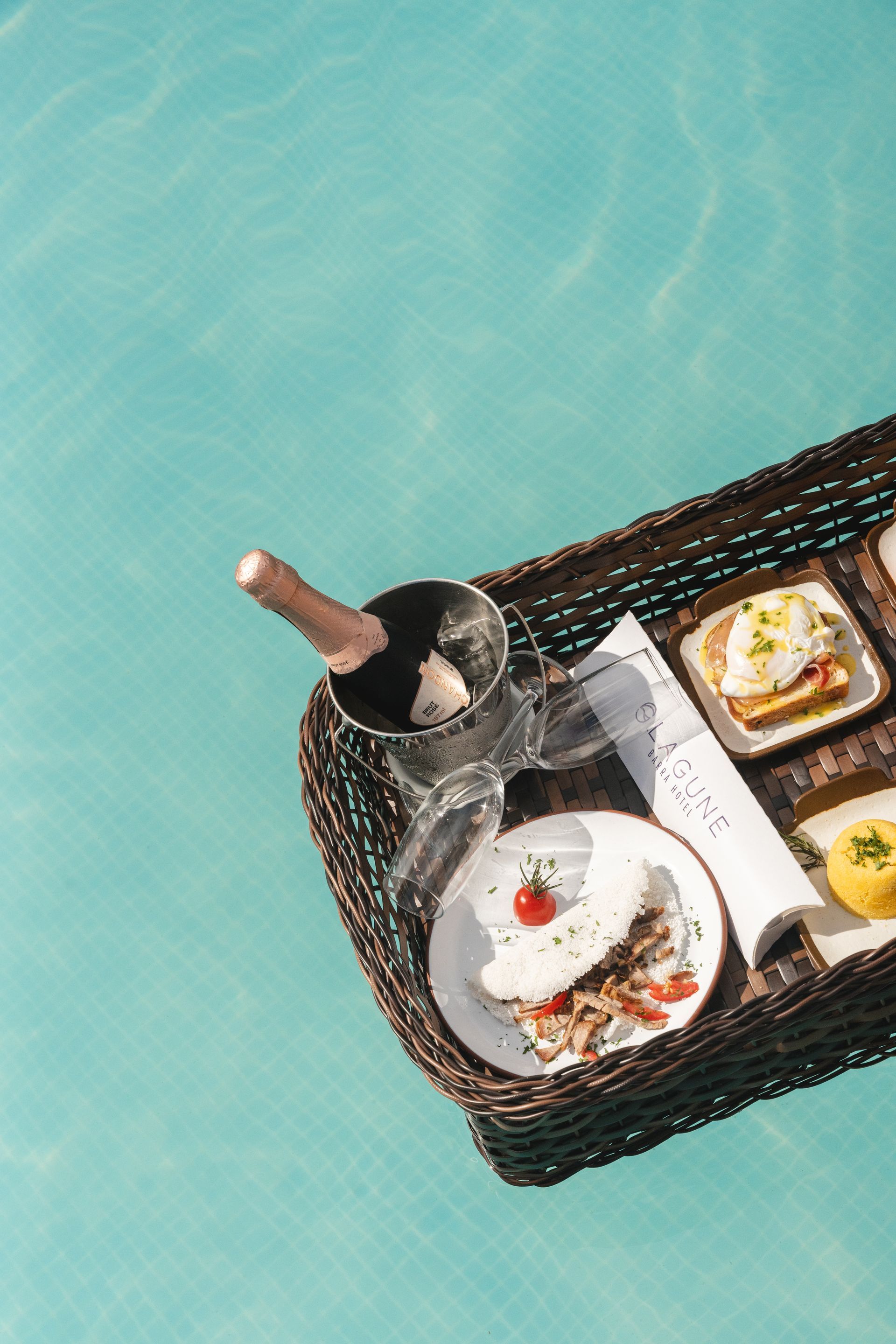 Bandeja de café da manhã flutuante em uma piscina: champanhe, ovos Benedict e um crepe, com água azul brilhante ao redor.