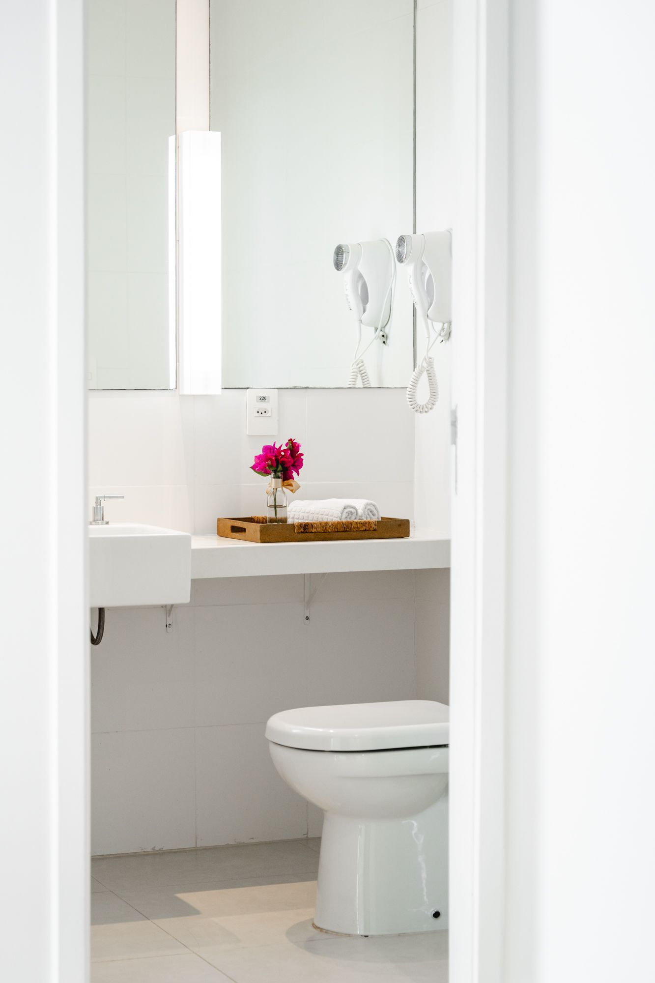 Banheiro branco brilhante visto de uma porta, com vaso sanitário, pia e balcão com flores e toalhas.