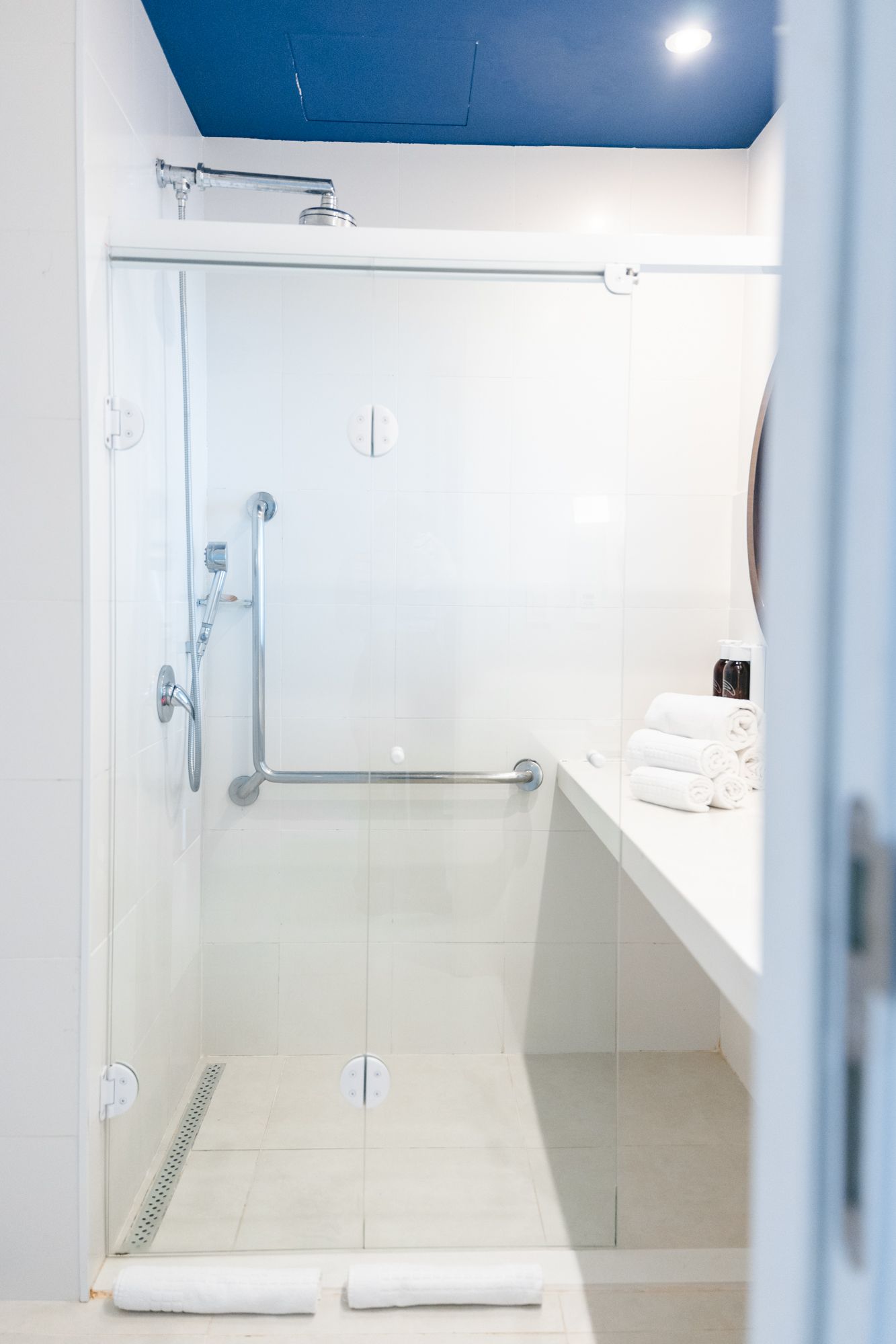 Banheiro branco acessível com box de vidro, corrimãos e uma pia com toalhas; teto azul.