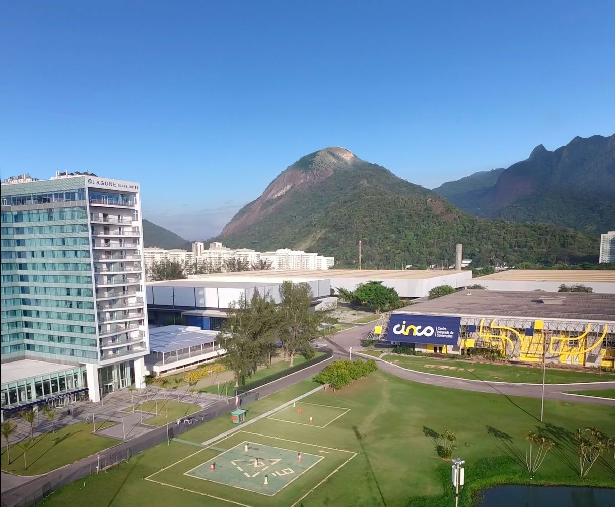 Prédio moderno e área verde em frente a uma montanha sob um céu azul. Ao longe, há outros prédios.