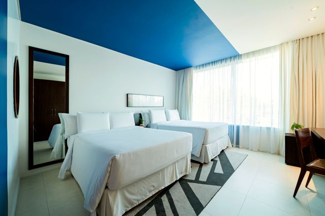 Duas camas de solteiro em um quarto de hotel moderno com detalhes em azul e branco, um espelho grande e uma janela com cortinas transparentes.