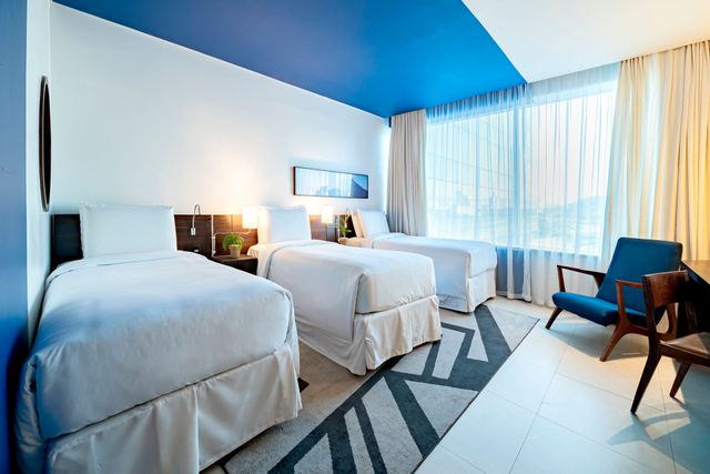 Um quarto de hotel com três camas de solteiro, uma poltrona azul e um tapete geométrico. O teto é parcialmente azul.