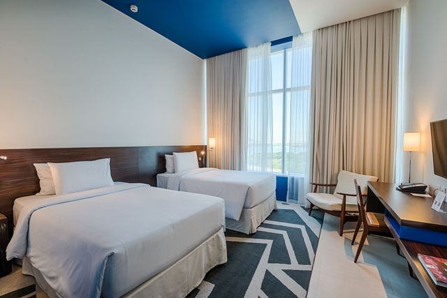 Quarto de hotel com duas camas, uma escrivaninha e uma cadeira perto de uma grande janela com cortinas transparentes; decoração em azul e branco.