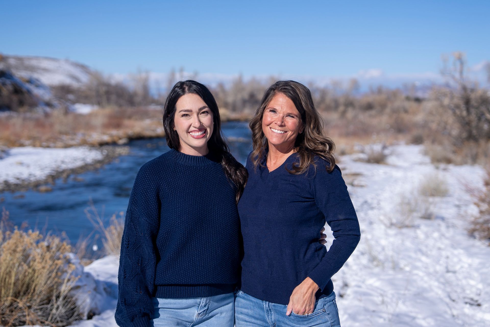 Two women stand smiling near a river, wearing matching blue sweaters, snow in background.