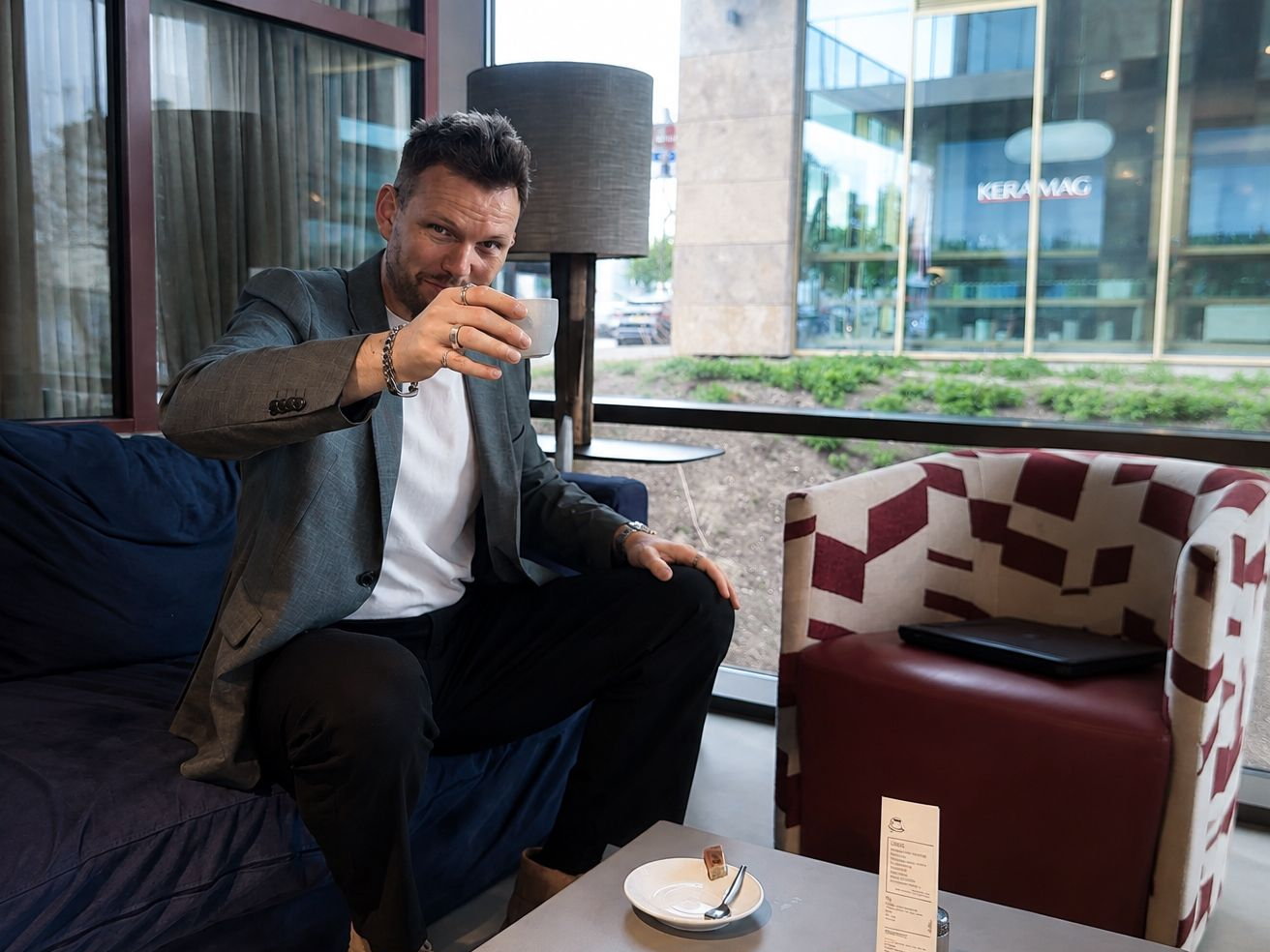 Man in a lounge points toward the camera while seated beside a table with dessert and drinks.