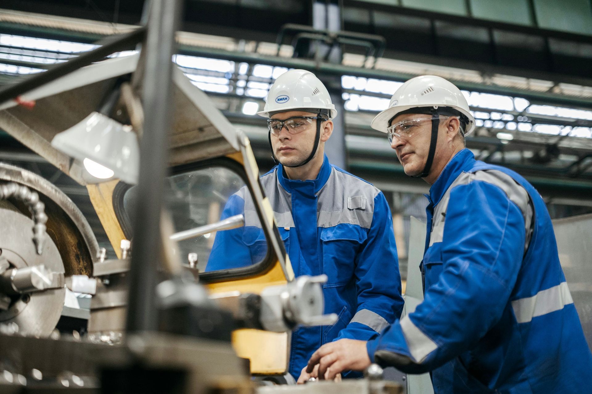 Two workers in blue uniforms and hard hats inspecting machinery in a factory.