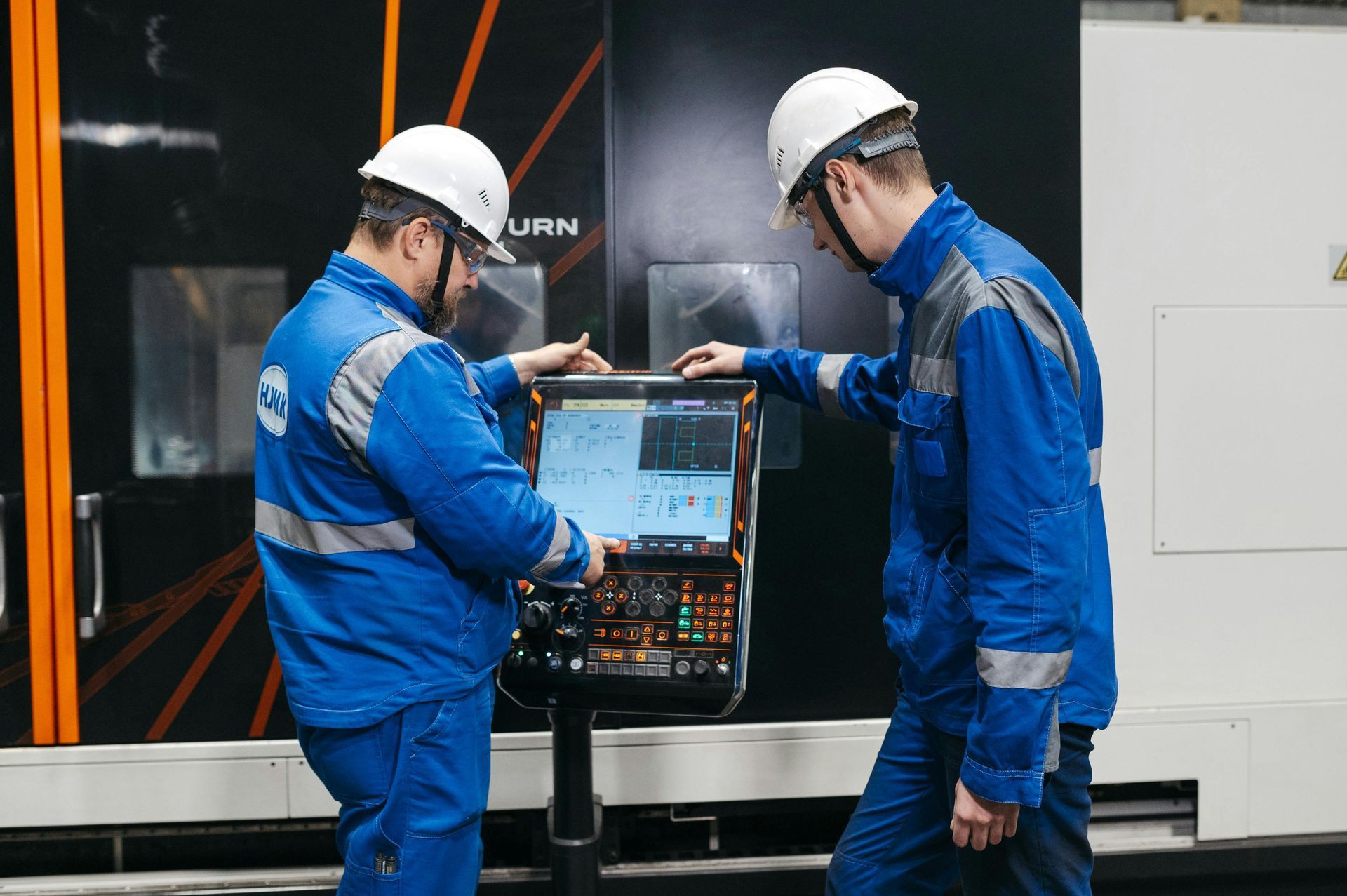 Two workers in blue coveralls inspect a machine in an industrial workshop.