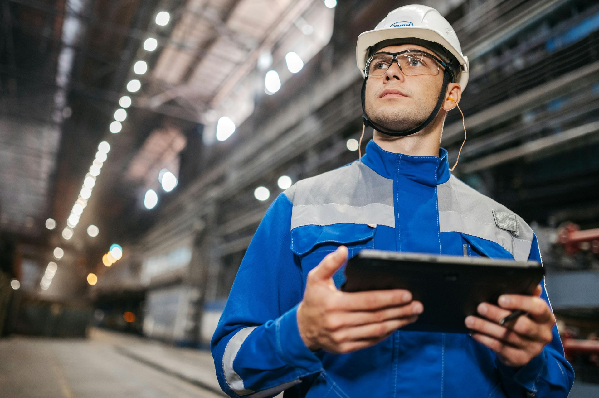 Industrial worker in blue uniform and hard hat holding a tablet in a factory aisle