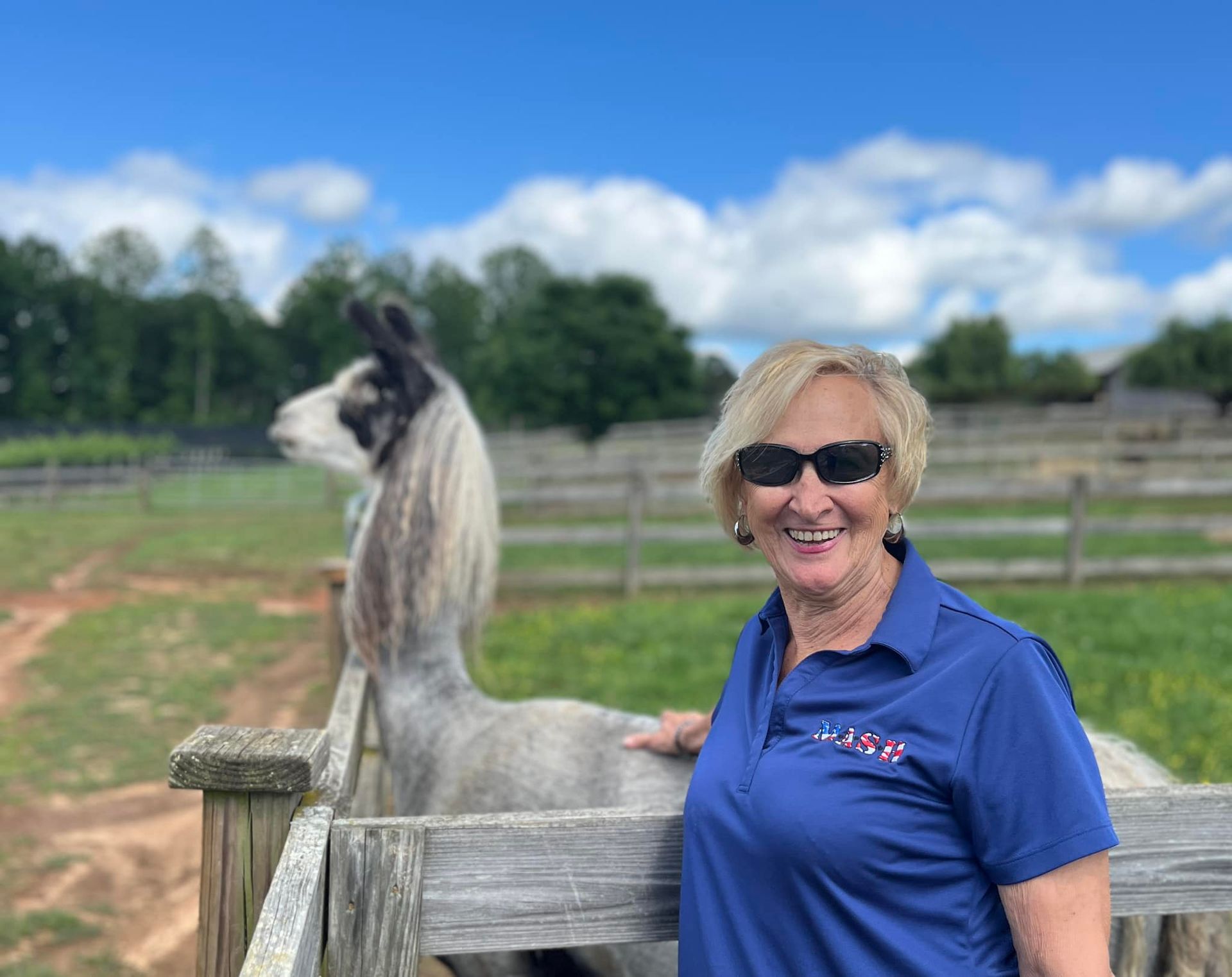 A woman is standing next to a fence with a llama in the background.
