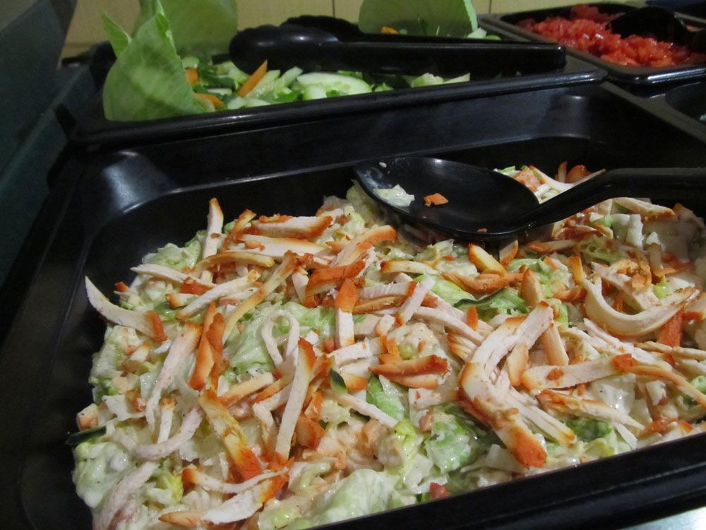 Salad bar with multiple trays of ingredients: lettuce, shredded chicken, cucumber and tomato.