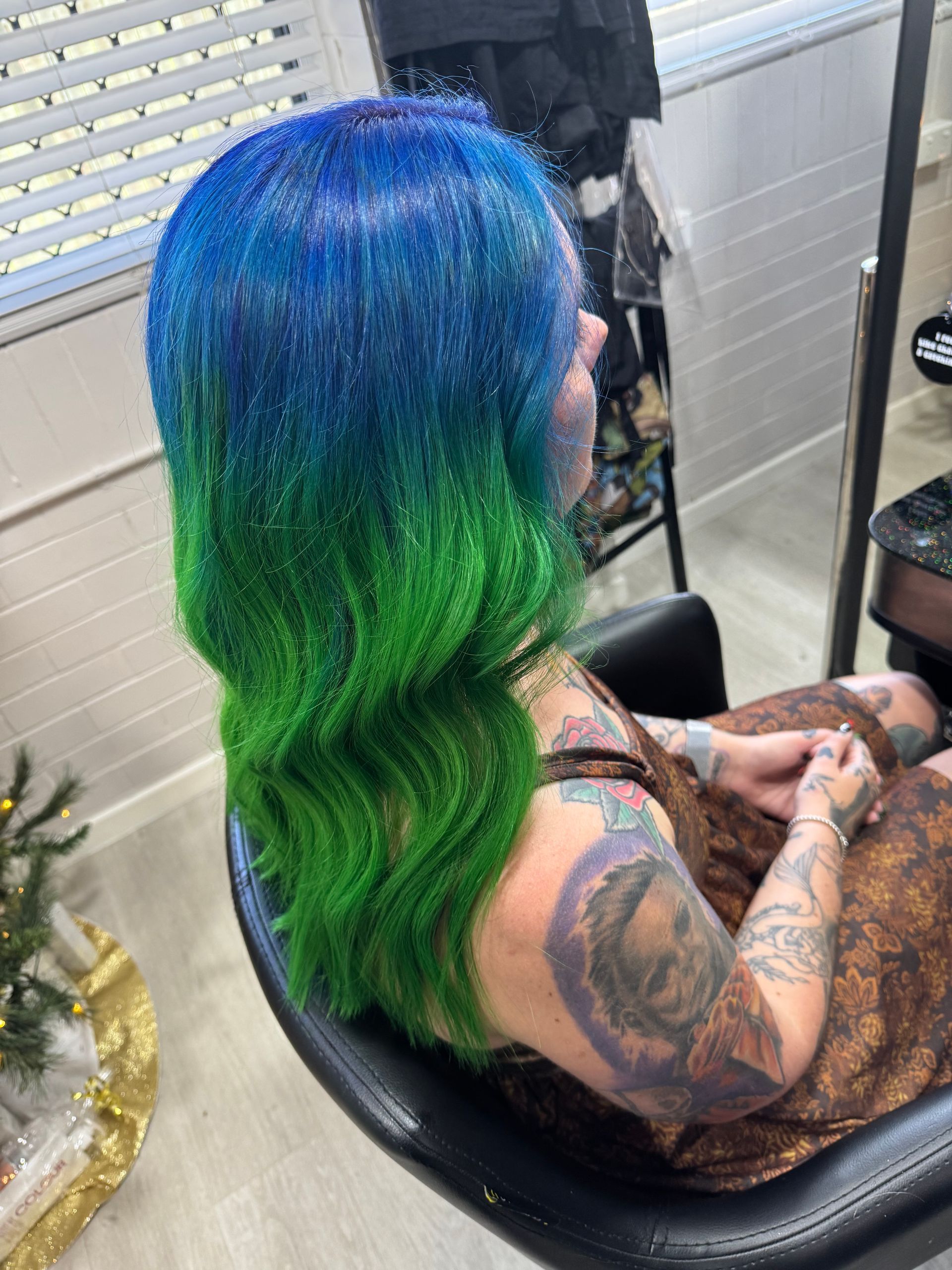 Woman with blue-to-green ombre hair sits in a salon chair. Her arm and shoulder are heavily tattooed.