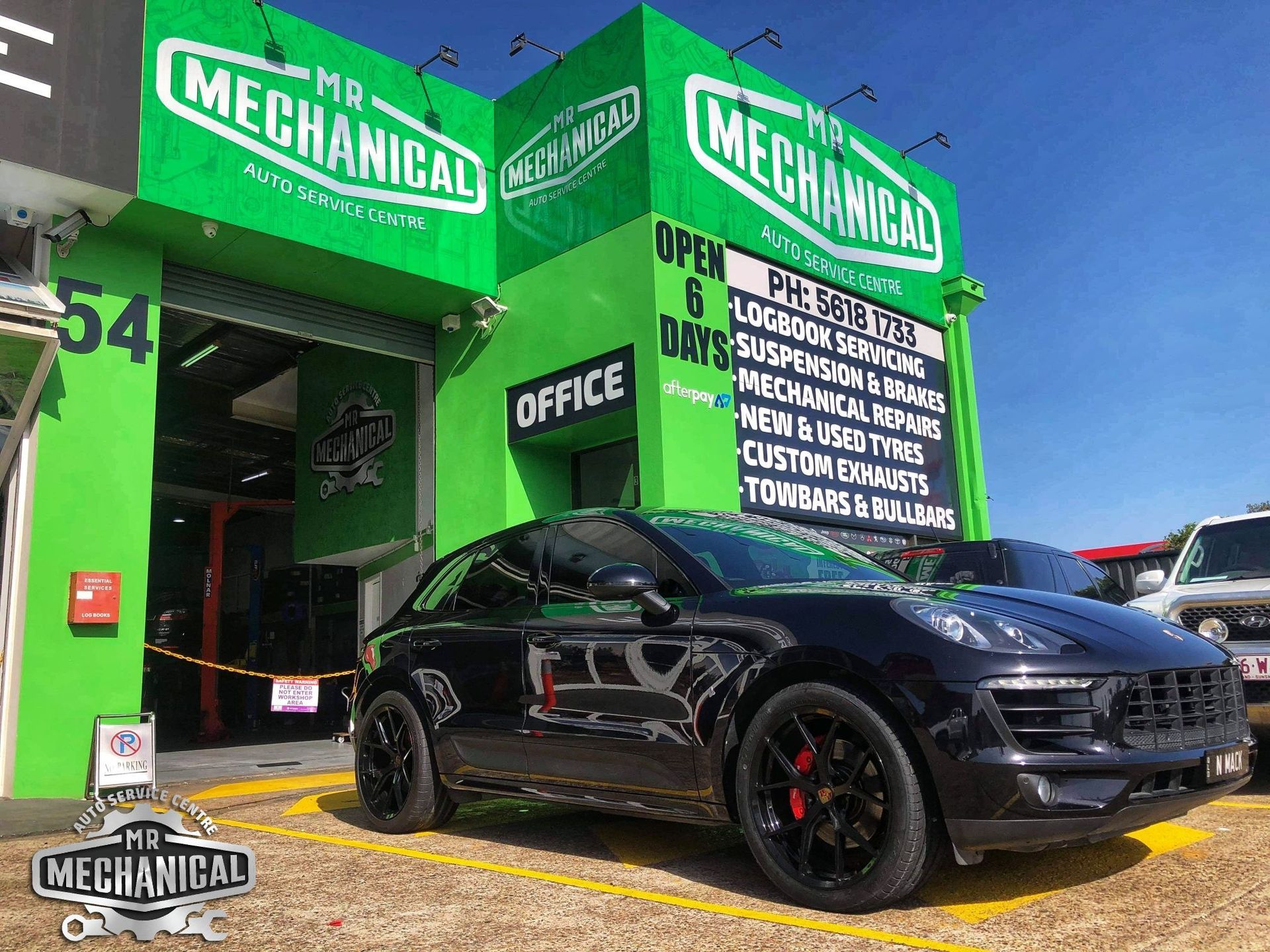 Black sports car parked outside a bright green MechaniKAL auto shop under a blue sky.