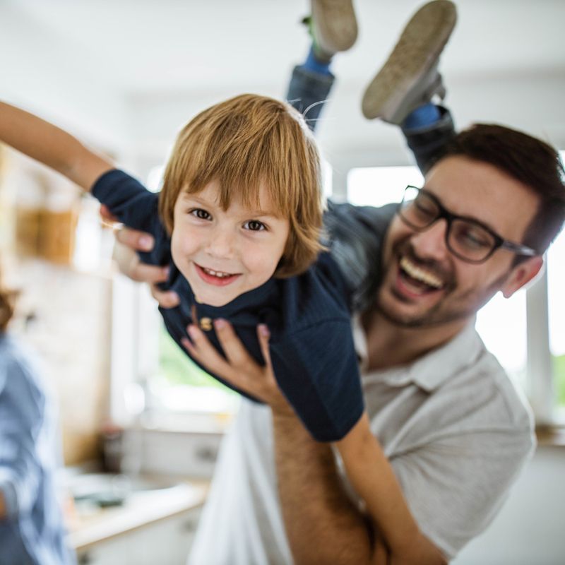 Smiling man playfully lifts a child indoors, both arms raised in a bright home setting