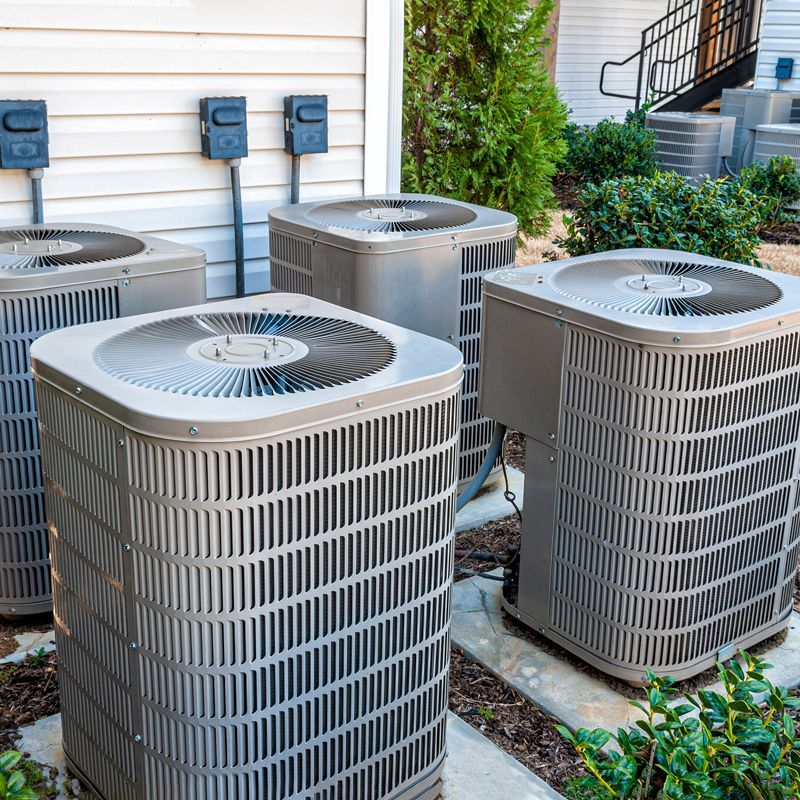 Four outdoor air conditioning units beside a house, with greenery and steps in the background