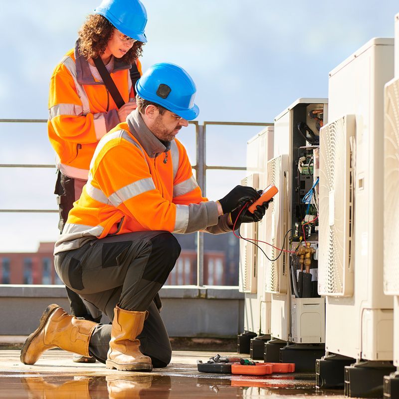 Two technicians in orange safety vests inspect electrical panels on a rooftop