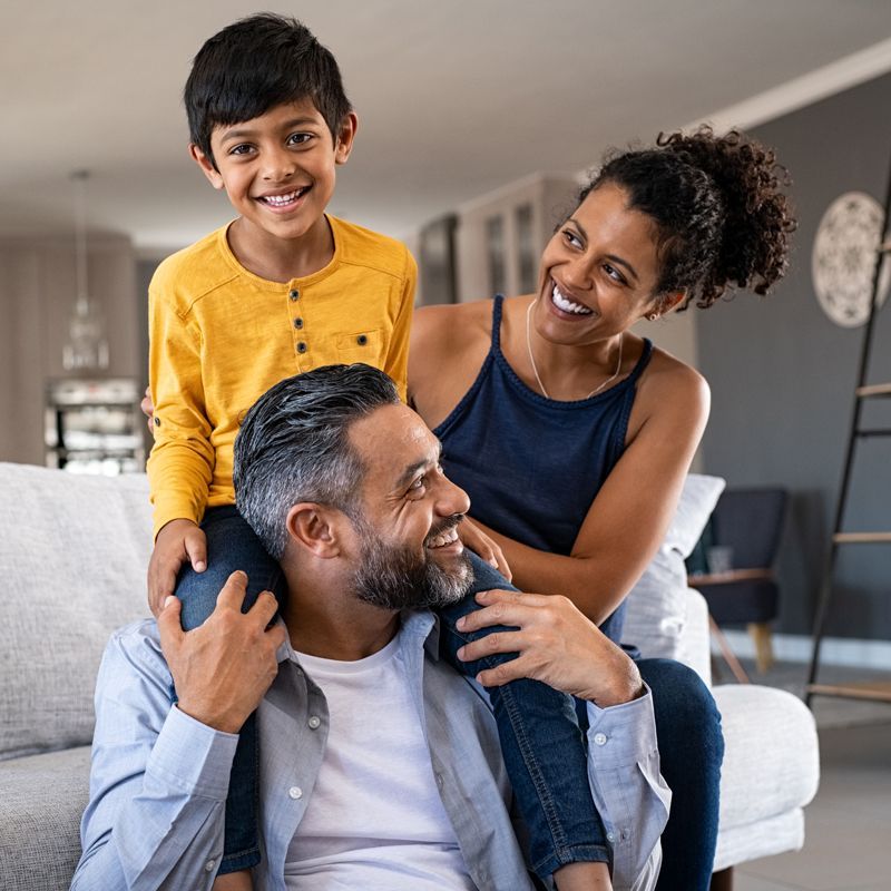 Smiling family indoors, with a child on a man's shoulders and a woman standing beside him.